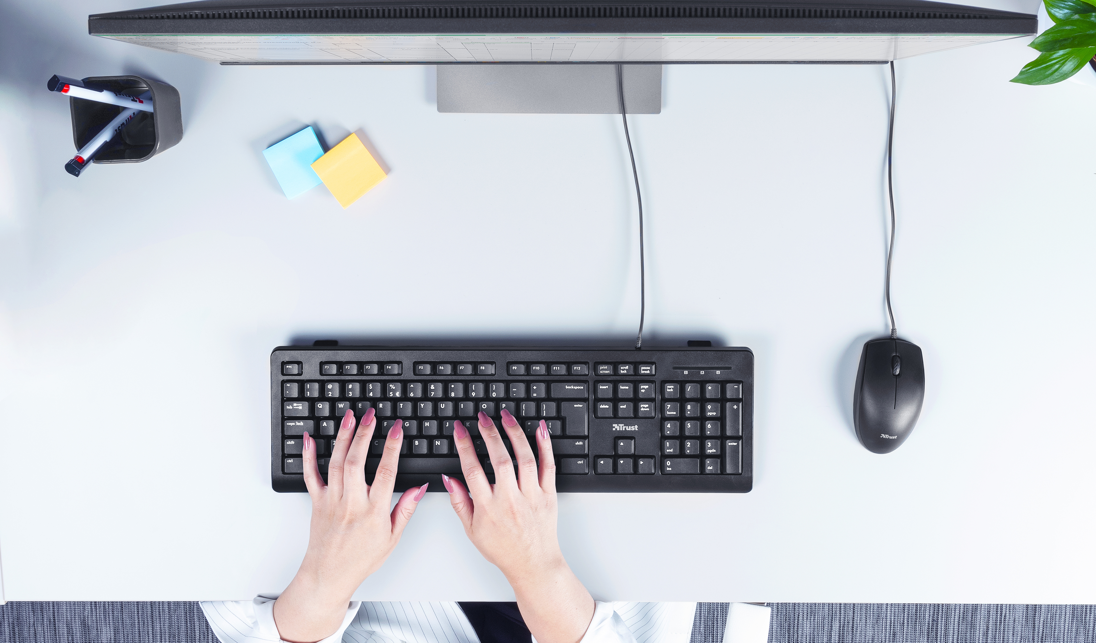 Widok z góry na ręce osoby piszącej na klawiaturze. Jest monitor, mysz i karteczki samoprzylepne.