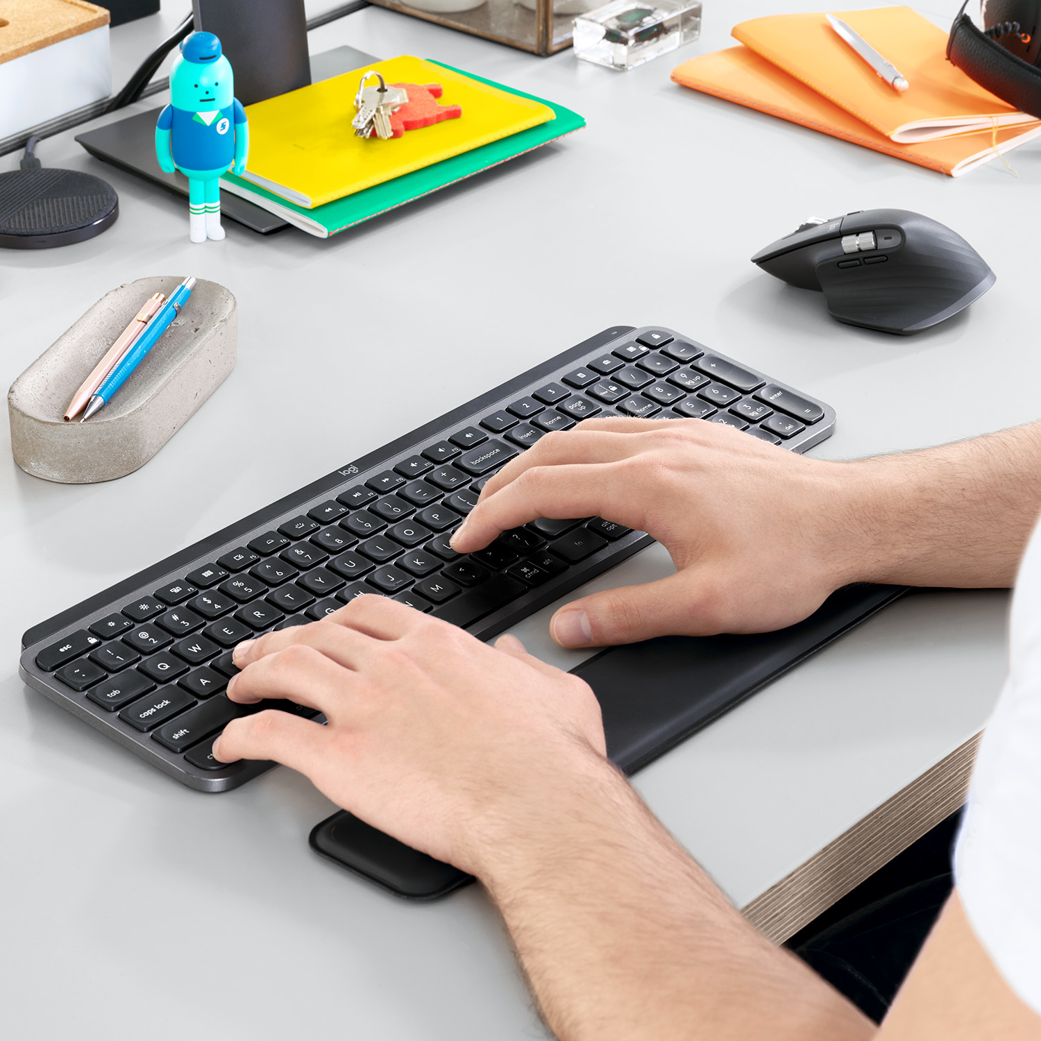 Mani che digitano su una tastiera con poggiapolsi. Scrivania con penne, mouse e piccole figure.