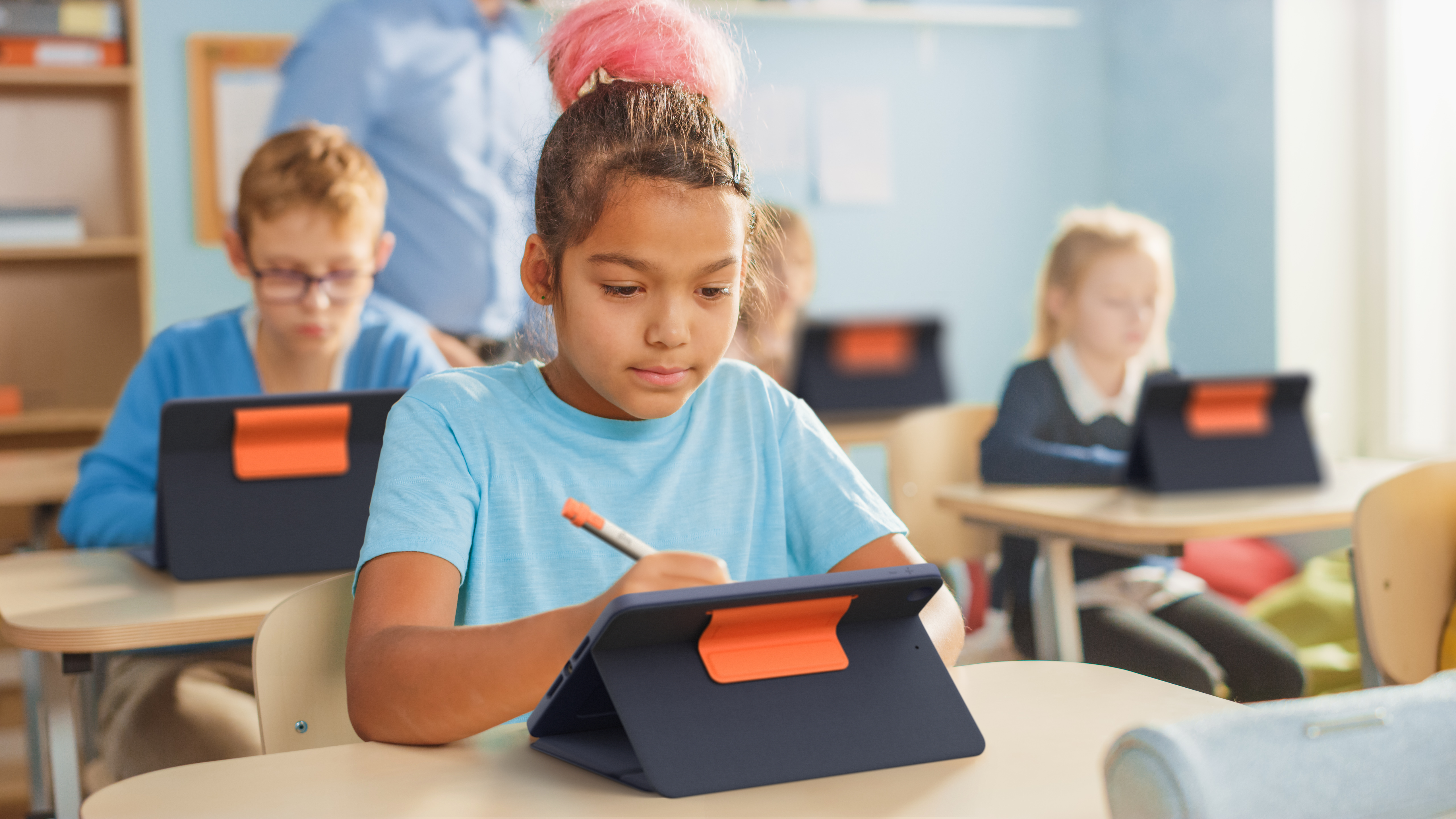 Ragazza con chignon rosa scrive su tablet in aula, insegnante sullo sfondo.