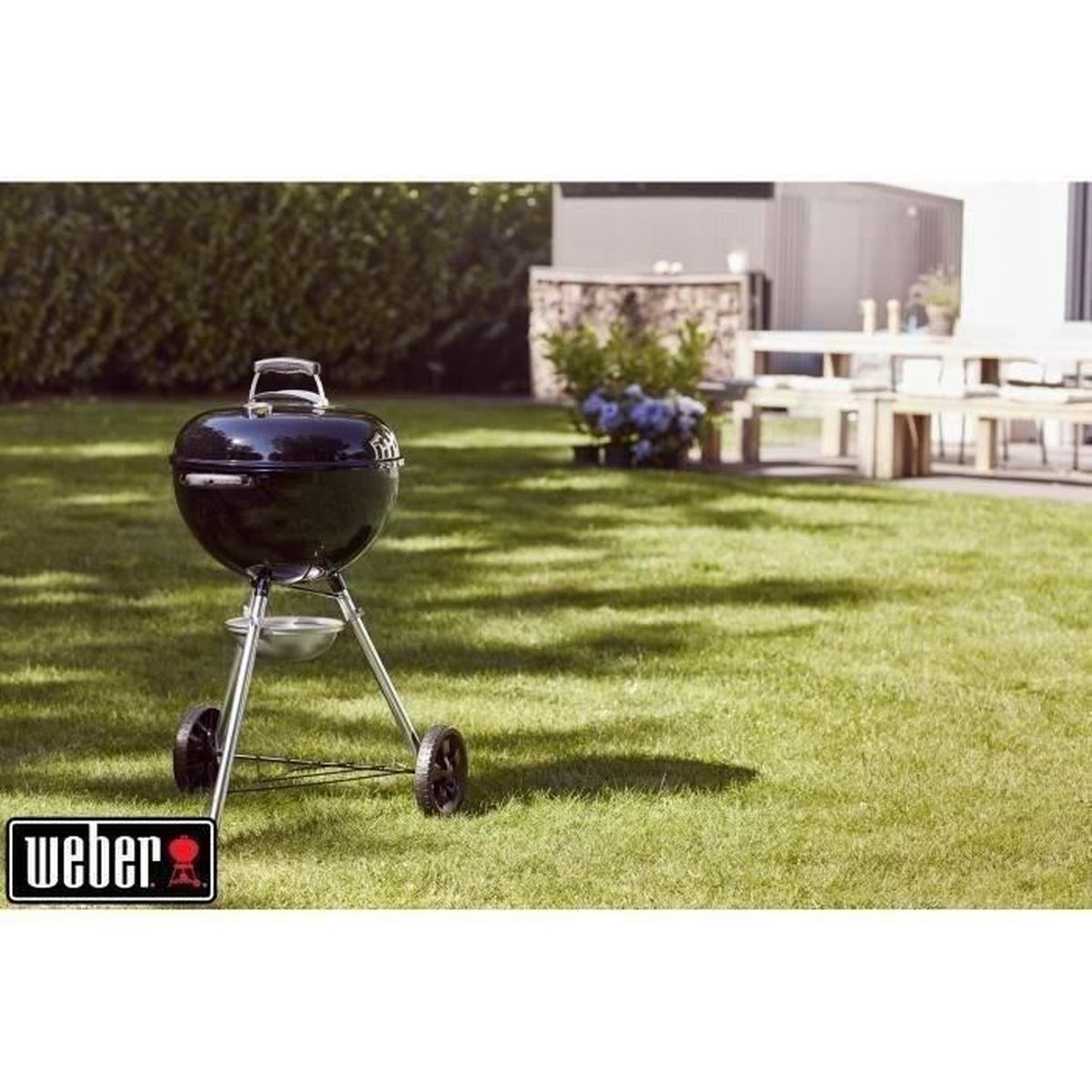 Czarny grill Weber na zielonym trawniku z domem i krzewami w tle.