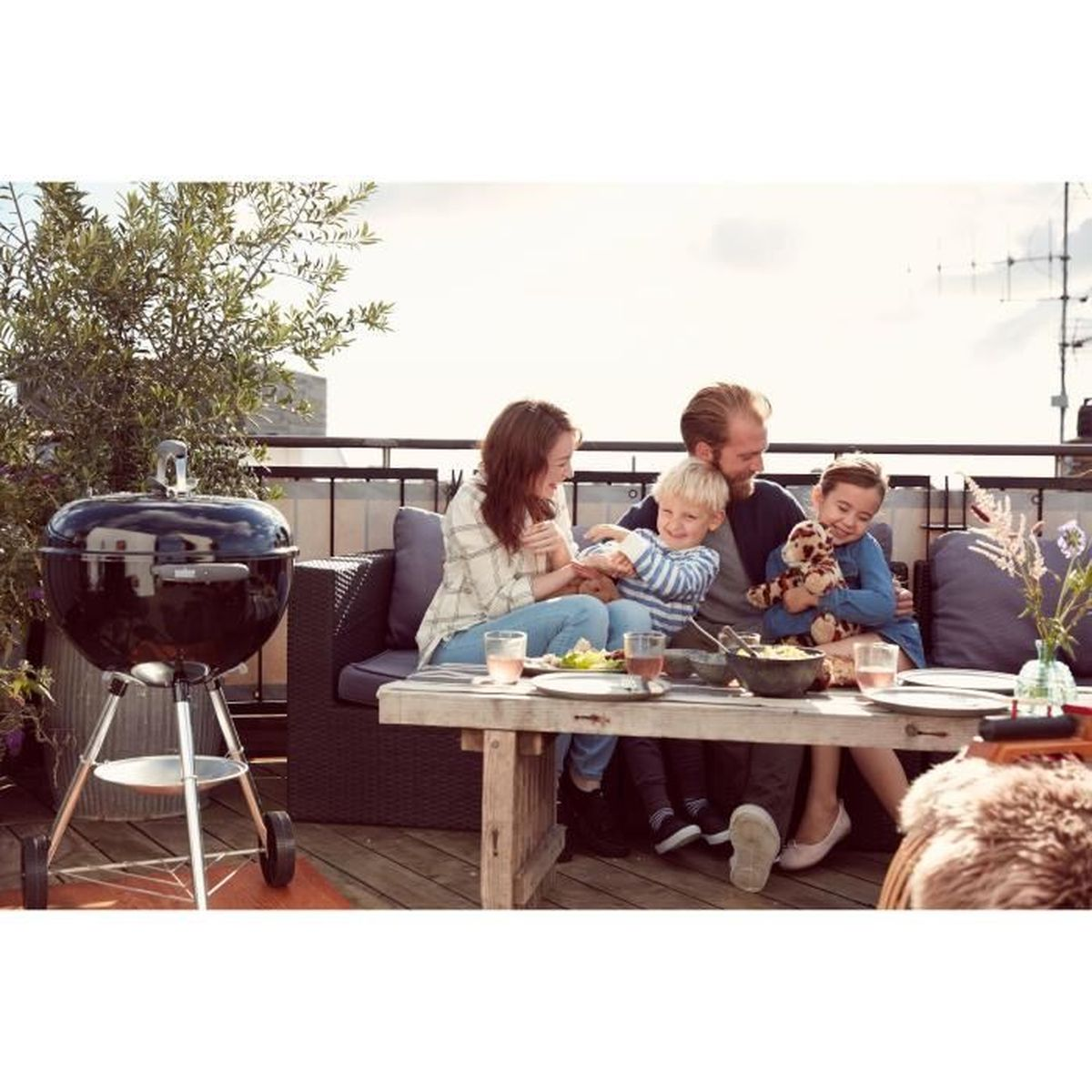 Famiglia ad un barbecue. Persone sedute a un tavolo, con un grill visibile. Ambiente esterno.
