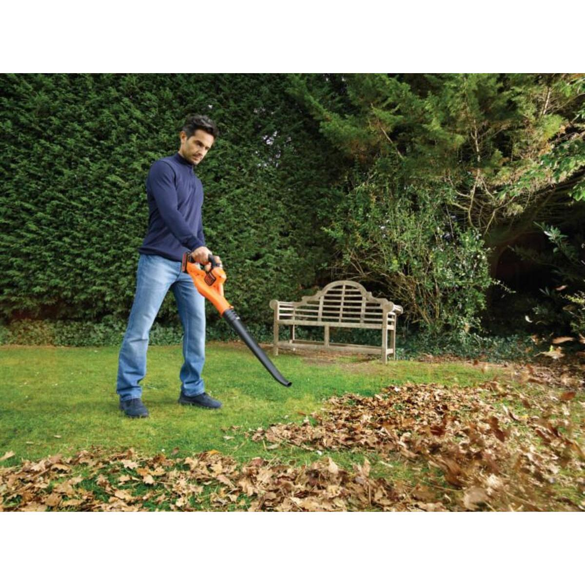 Man blaast bladeren met een oranje bladblazer. Hij is in een tuin met een haag en een bank.