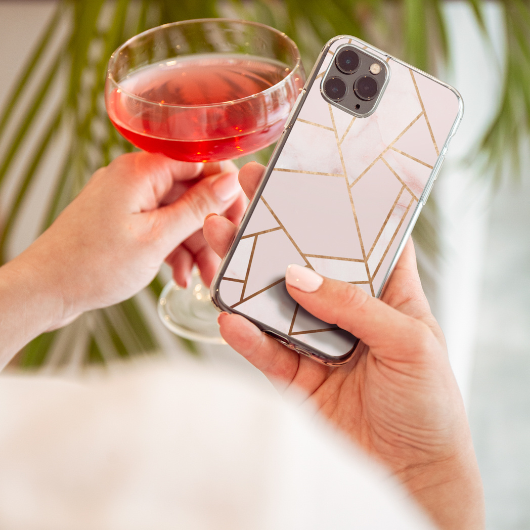 Persoon houdt telefoon vast in een doorzichtige hoes met geometrisch goud design, bij een drankje.