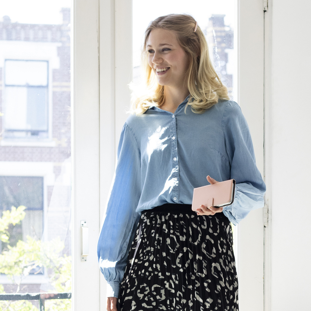 Een lachende vrouw met blond haar houdt een roze telefoonhoesje voor een raam.