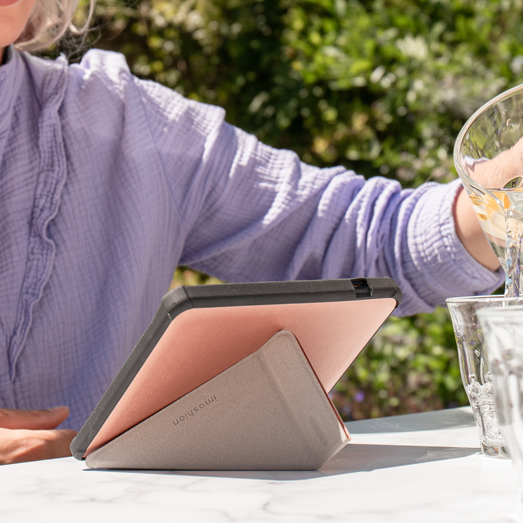 Personne versant de l'eau dans un verre, tablette avec étui, sur une table à l'extérieur, ensoleillé.