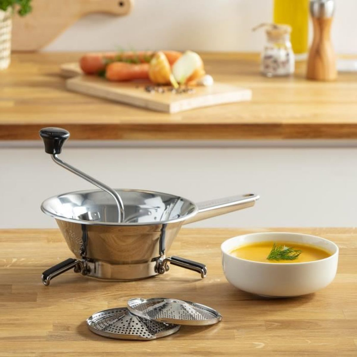 Eine Edelstahl-Lebensmittelmühle, Karotten, Zwiebeln und eine Schüssel Suppe auf einem Holztisch.