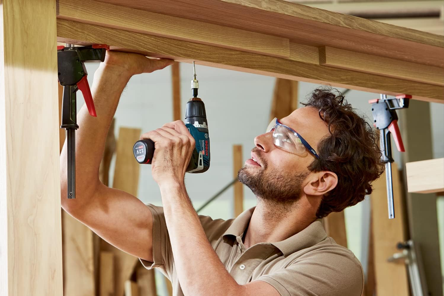 Uomo usa trapano su tavole di legno. Indossa occhiali di sicurezza, concentrato.