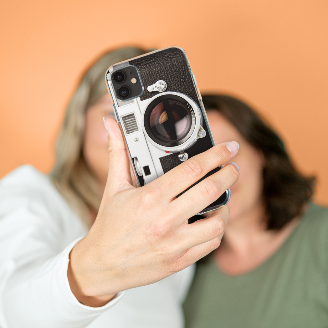 Twee vrouwen nemen een selfie met een telefoonhoesje dat eruitziet als een camera. Oranje achtergrond.