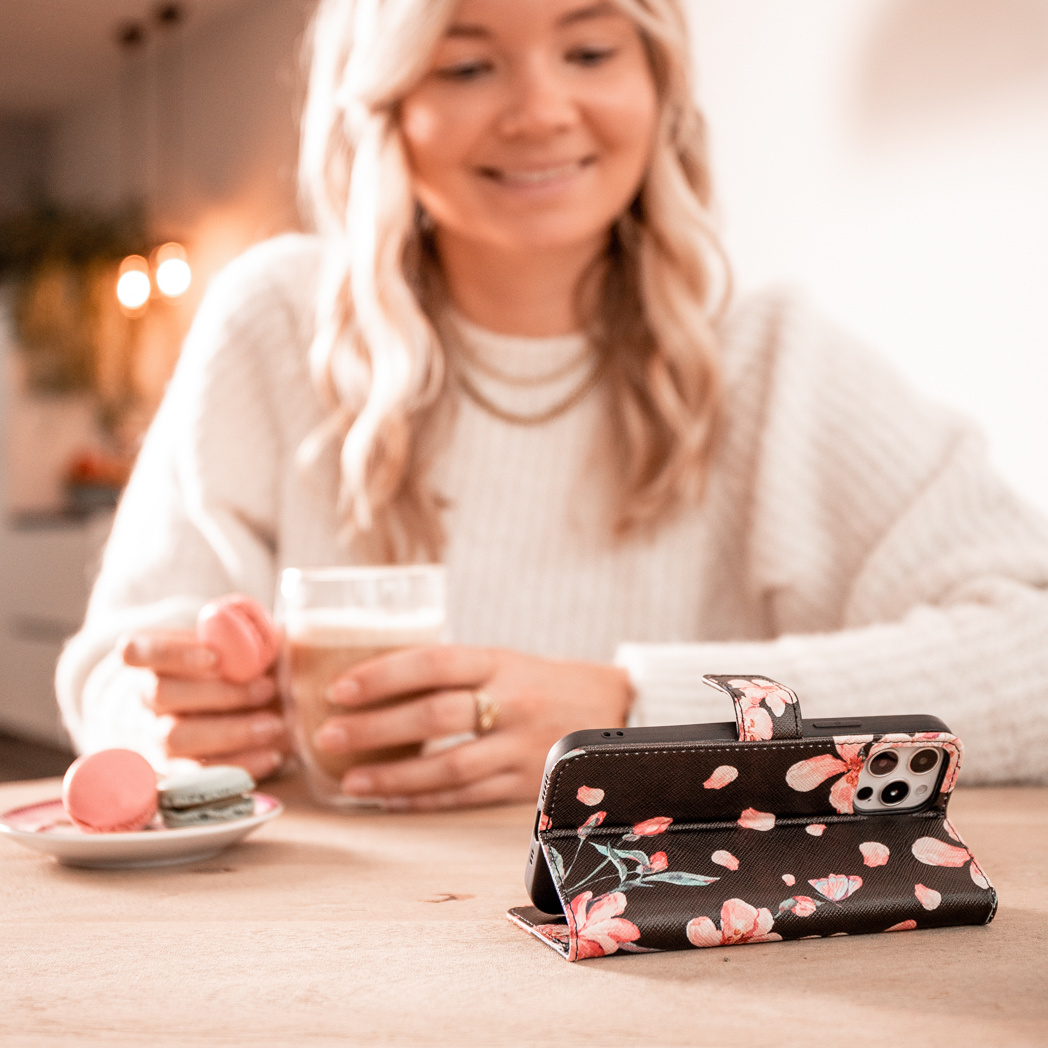 Vrouw glimlachend aan een tafel met een telefoonhoesje met bloemen open, en een drankje voor haar.