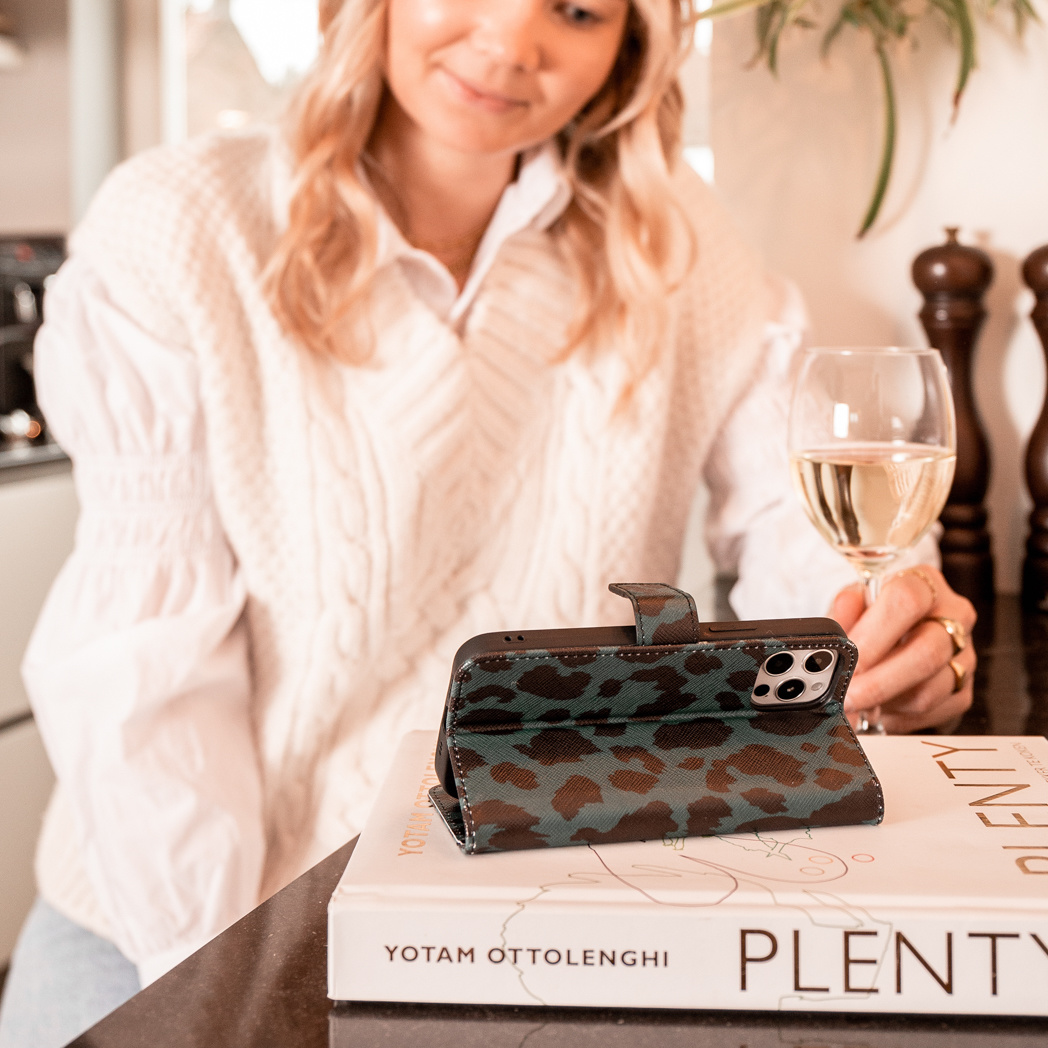 Vrouw die een telefoon in een hoesje vasthoudt. Boek en wijnglas op tafel.