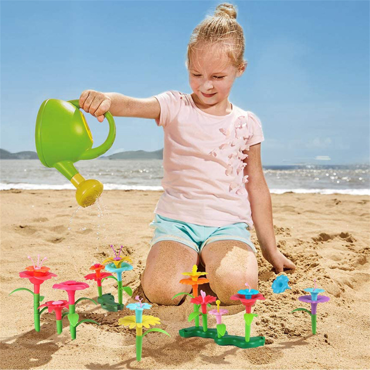 Ein Mädchen gießt Spielzeugblumen am Strand. Blauer Himmel und Sand im Hintergrund.