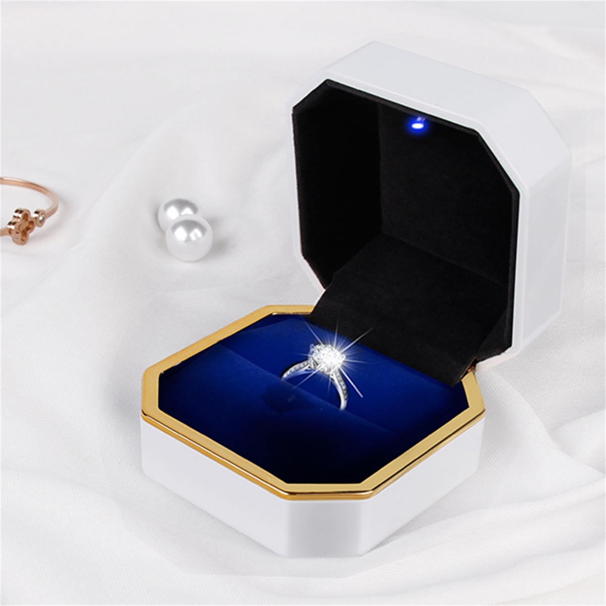Eine weiße Schmuckschatulle mit einem Diamantring darin, auf weißer Oberfläche.