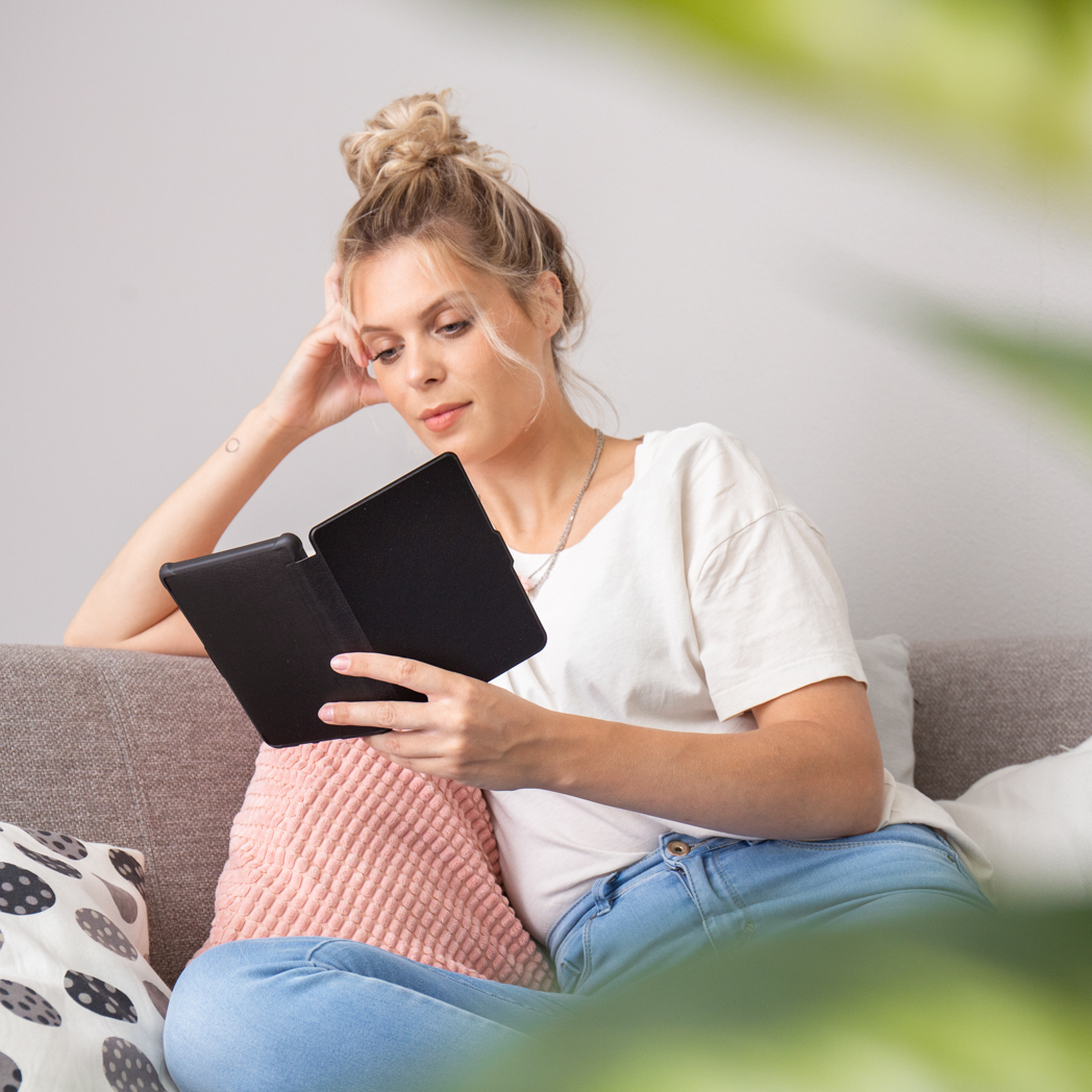 Une femme lisant un lecteur électronique noir sur un canapé. Elle est appuyée, avec un coussin rose et une plante verte.