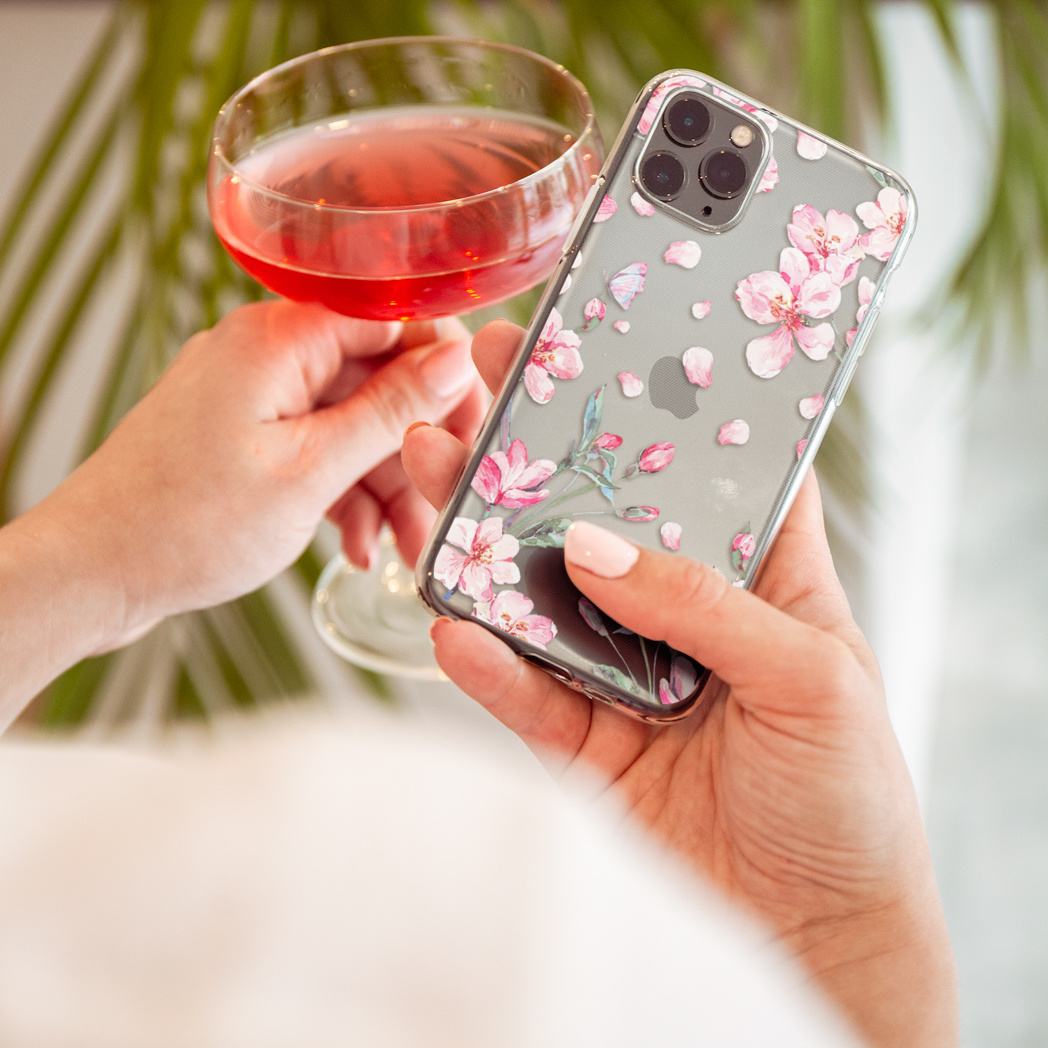 Een persoon houdt een telefoon met een bloemenhoesje en een glas roze vloeistof vast.