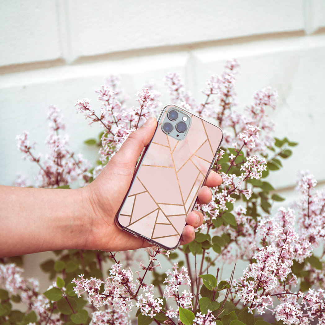 Main tenant un téléphone avec un étui au design géométrique rose et doré, entouré de fleurs roses et de feuilles vertes.