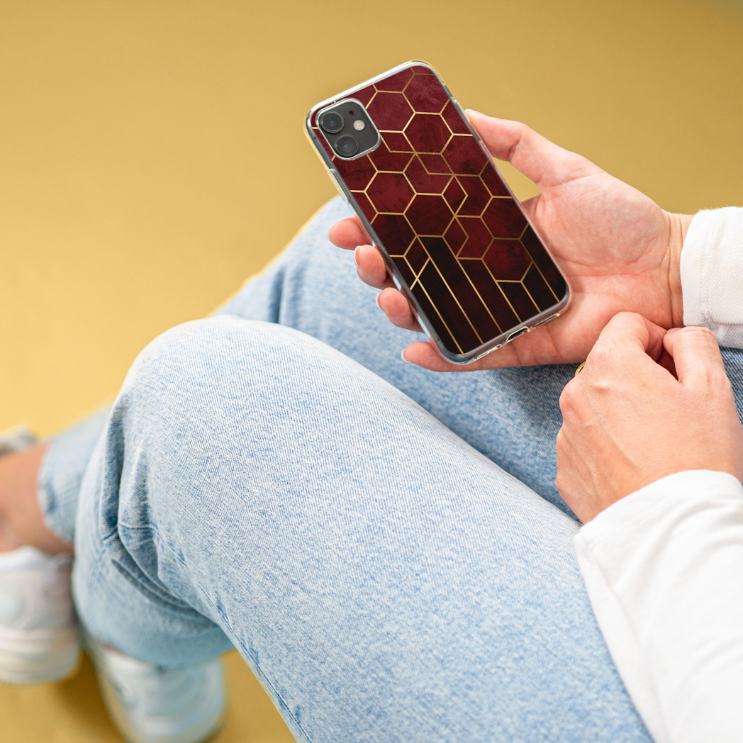 Persoon die een telefoon vasthoudt met een bordeauxrode en gouden geometrische hoes. Gele achtergrond.