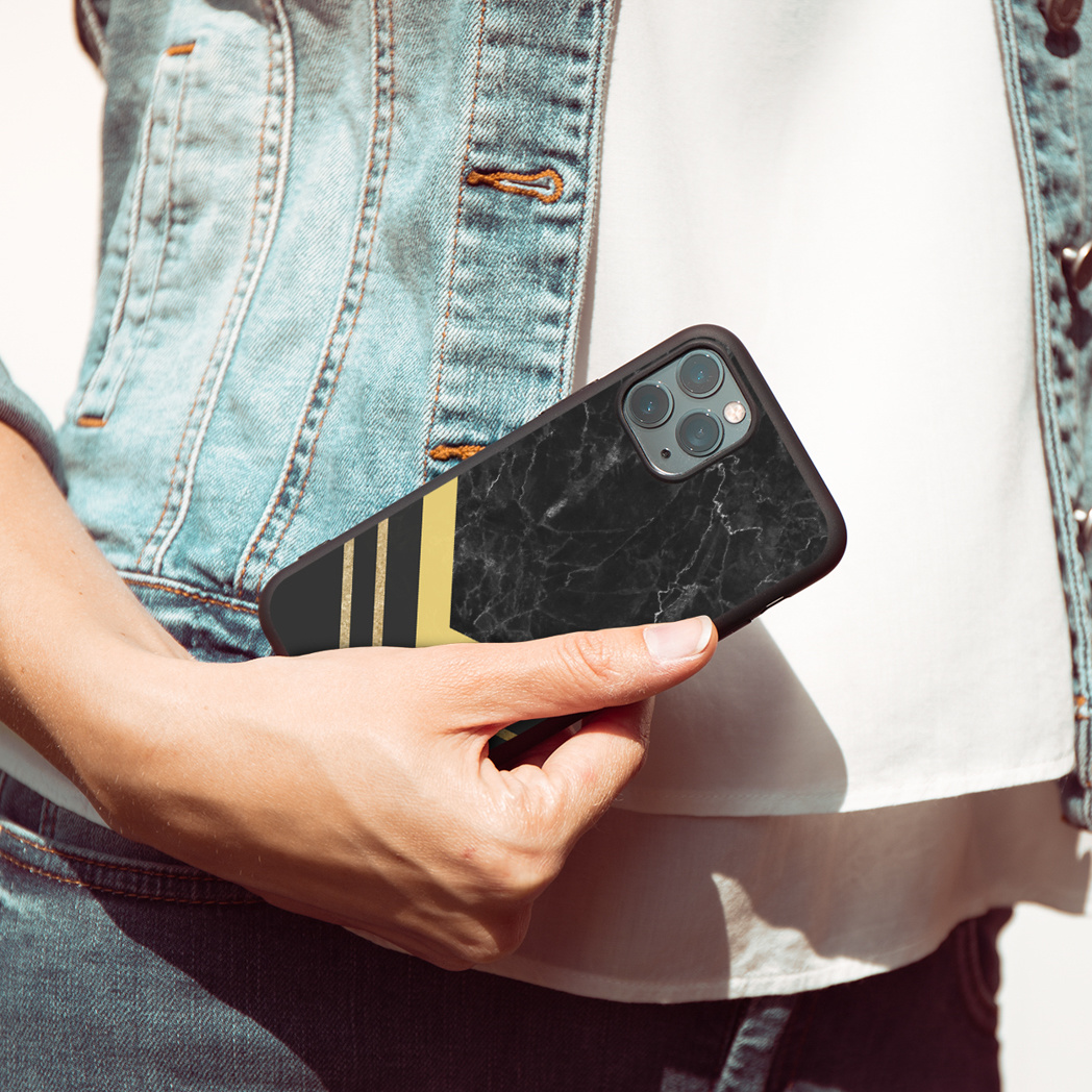 Een persoon die een zwarte telefoonhoes vasthoudt met een marmerdesign en gouden strepen, tegen een spijkerjasje.