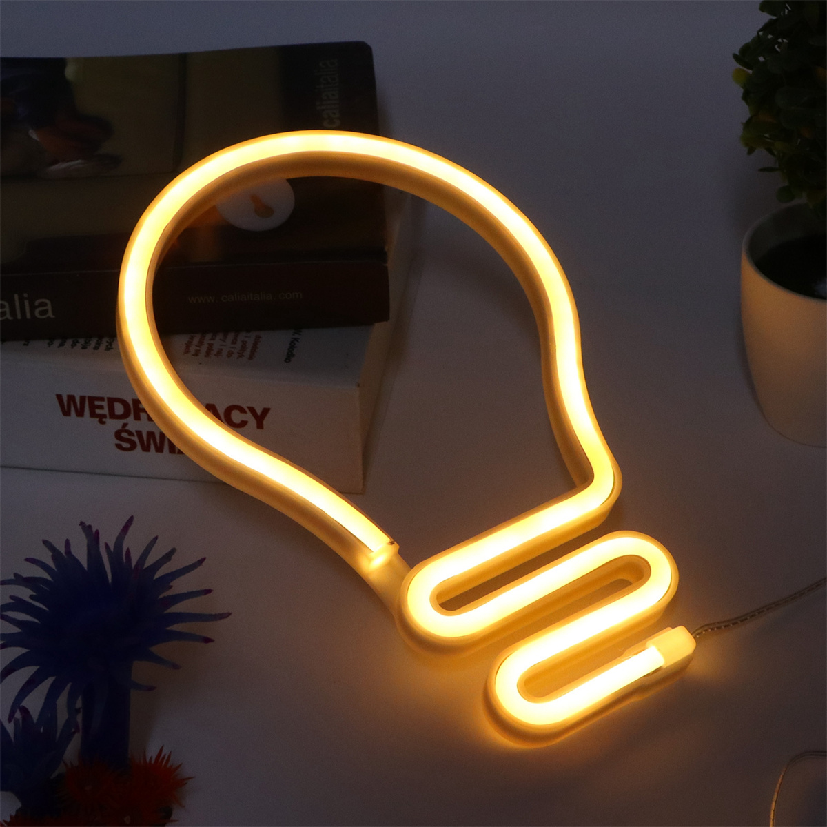 Gelbes Neonlicht in Form einer Glühbirne auf weißer Oberfläche, mit Büchern und einer Pflanze im Hintergrund.
