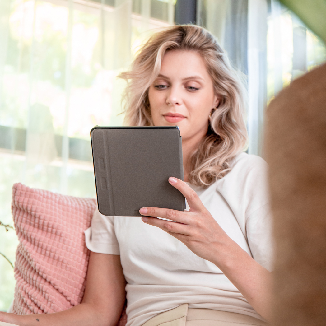 Vrouw houdt een grijze tablet vast. Zittend voor een raam. Vage achtergrond.