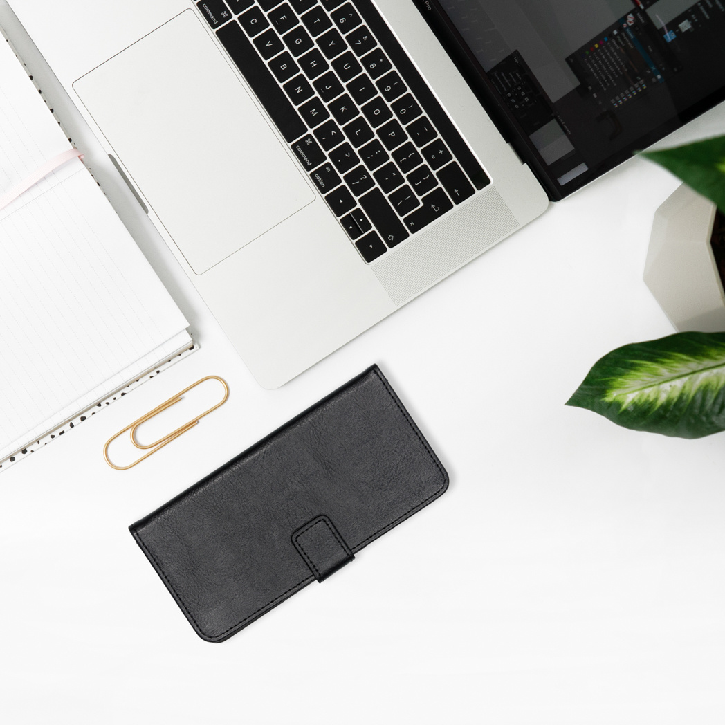 Un étui de téléphone noir et des accessoires sur un bureau blanc. Un ordinateur portable et une plante en arrière-plan.