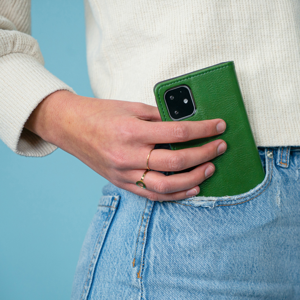 Hand houdt een groene telefoonhoes in een zak. Spijkerbroek en een witte trui zijn zichtbaar.
