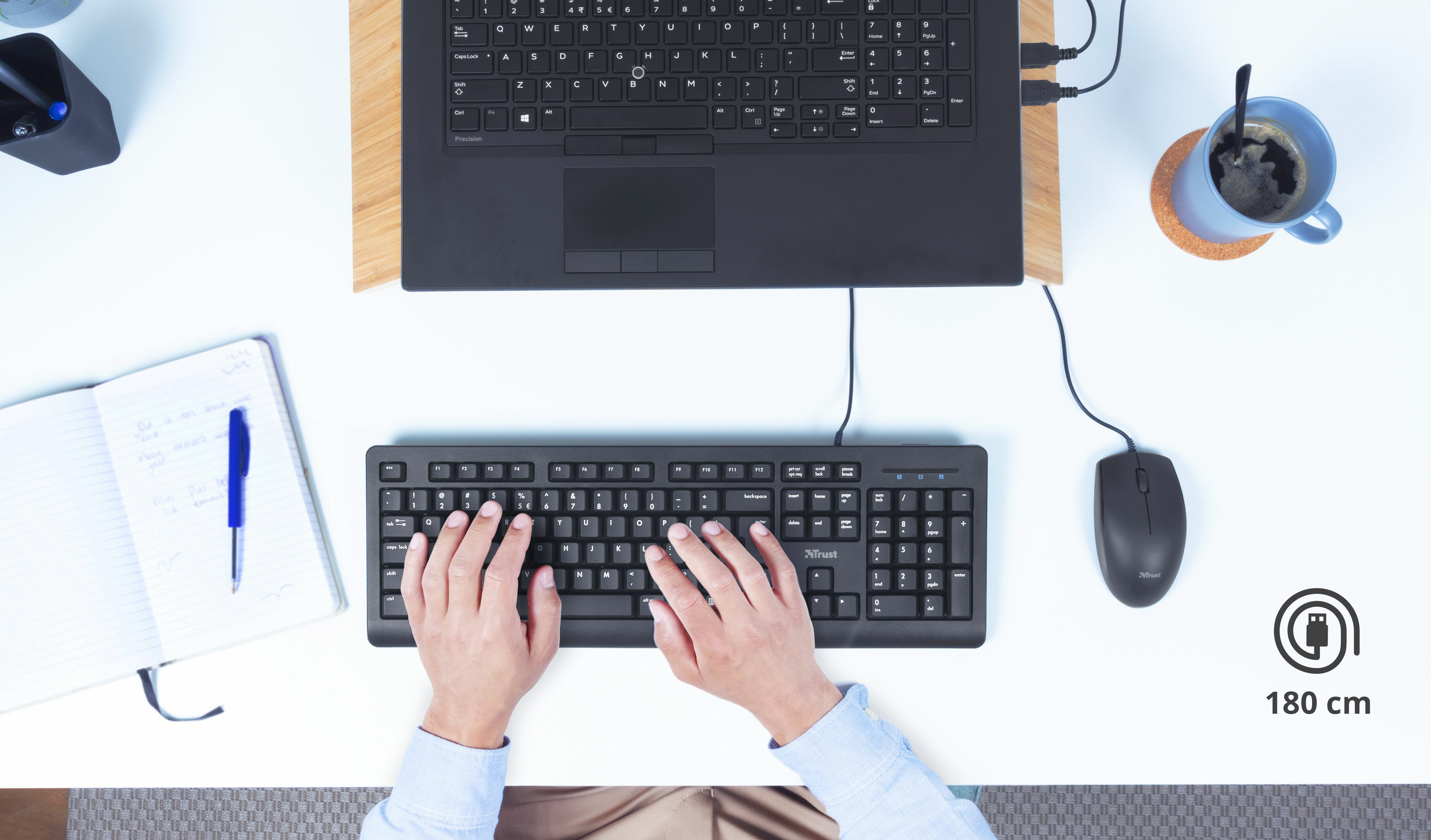 Osoba pisząca na klawiaturze. Na biurku znajduje się również laptop, długopis i filiżanka kawy.