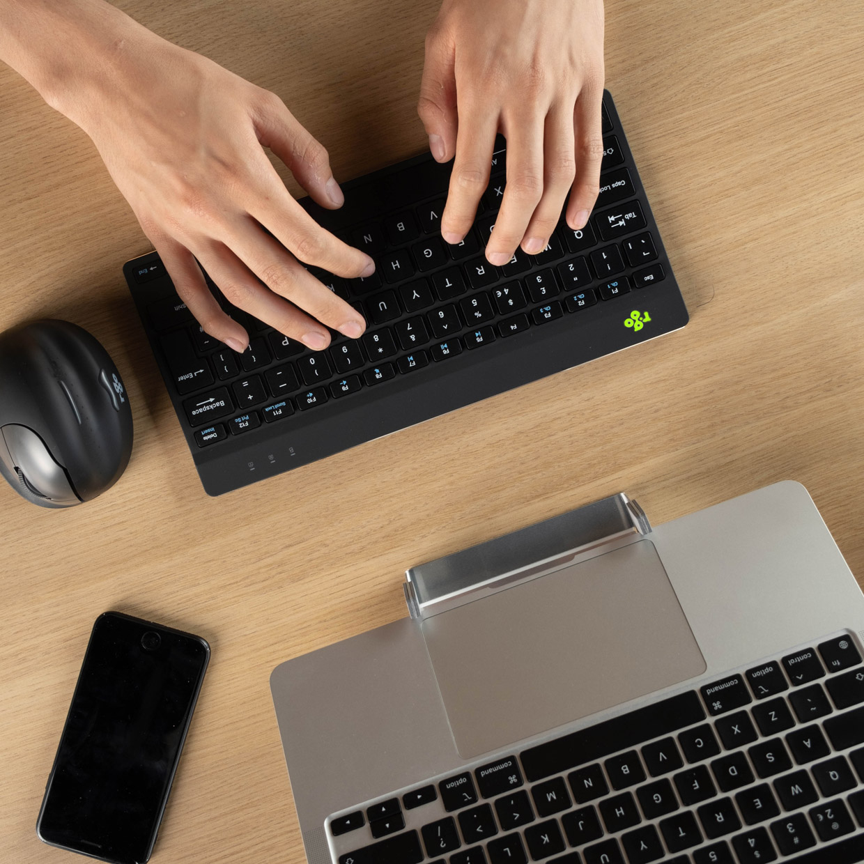 Hände tippen auf einer schwarzen Tastatur auf einem Holztisch. Eine Maus und ein Laptop sind ebenfalls sichtbar.