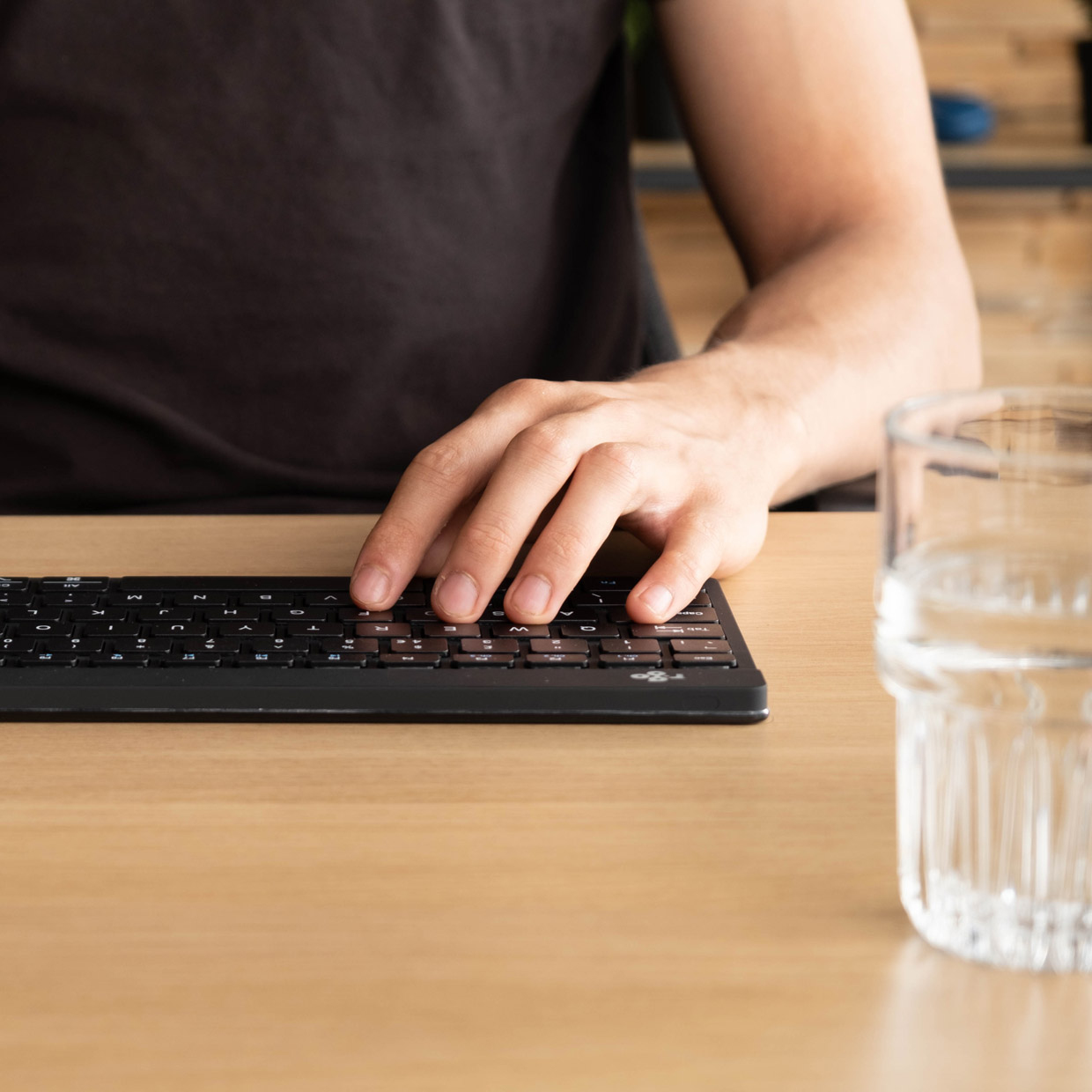 Hände einer Person, die auf einer schwarzen Tastatur tippen. Rechts ein Glas Wasser.