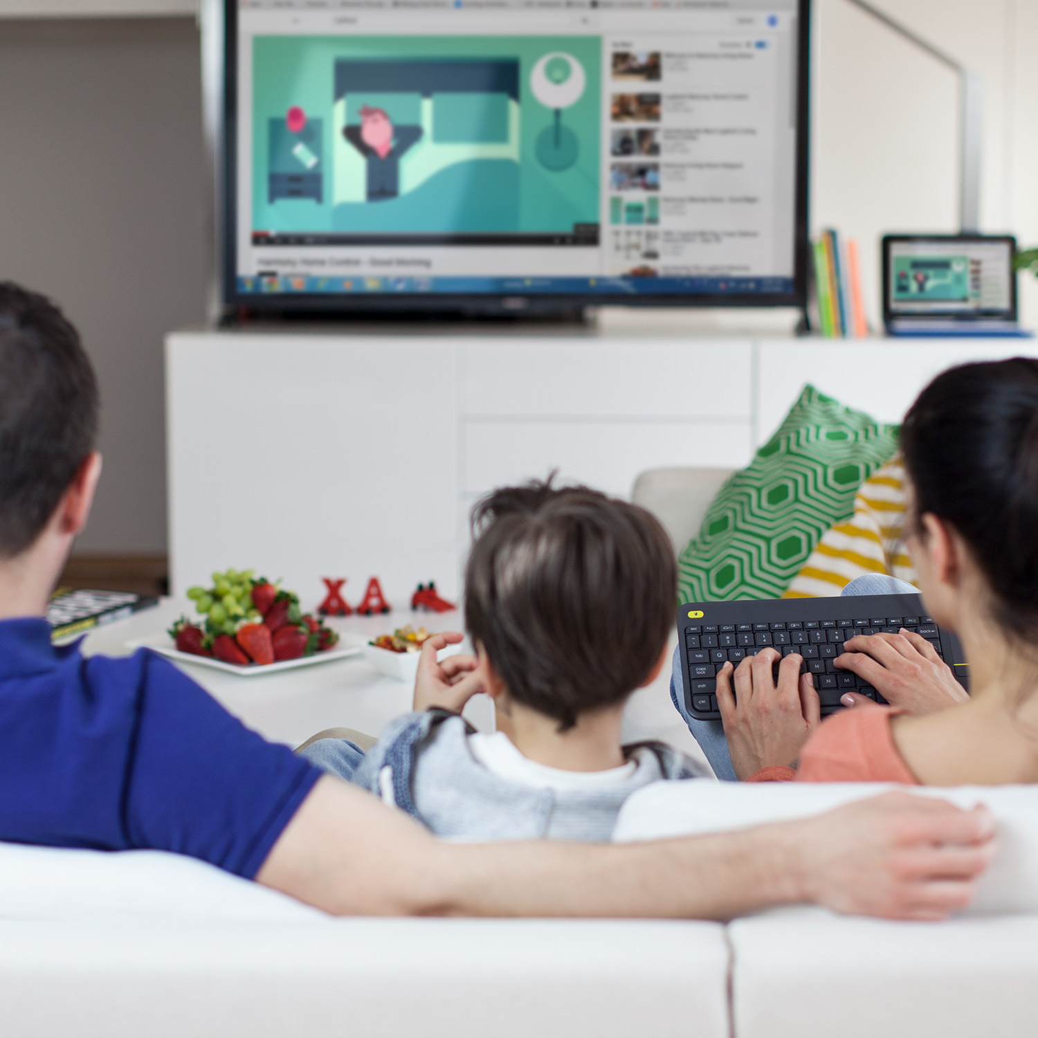 Rodzina ogląda telewizję, z klawiaturą na pierwszym planie. Dziecko siedzi na kanapie.