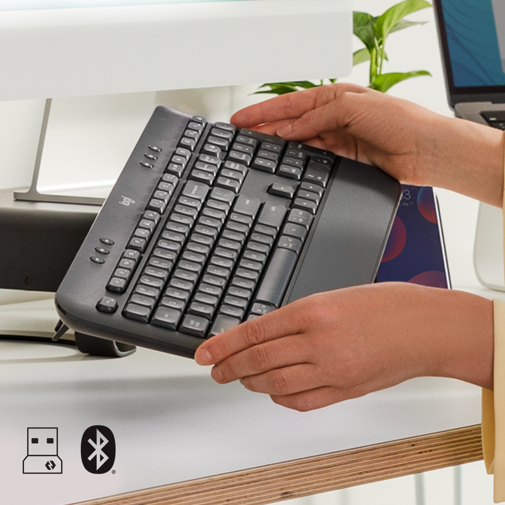 Dłonie trzymające czarną ergonomiczną klawiaturę nad białym biurkiem.
