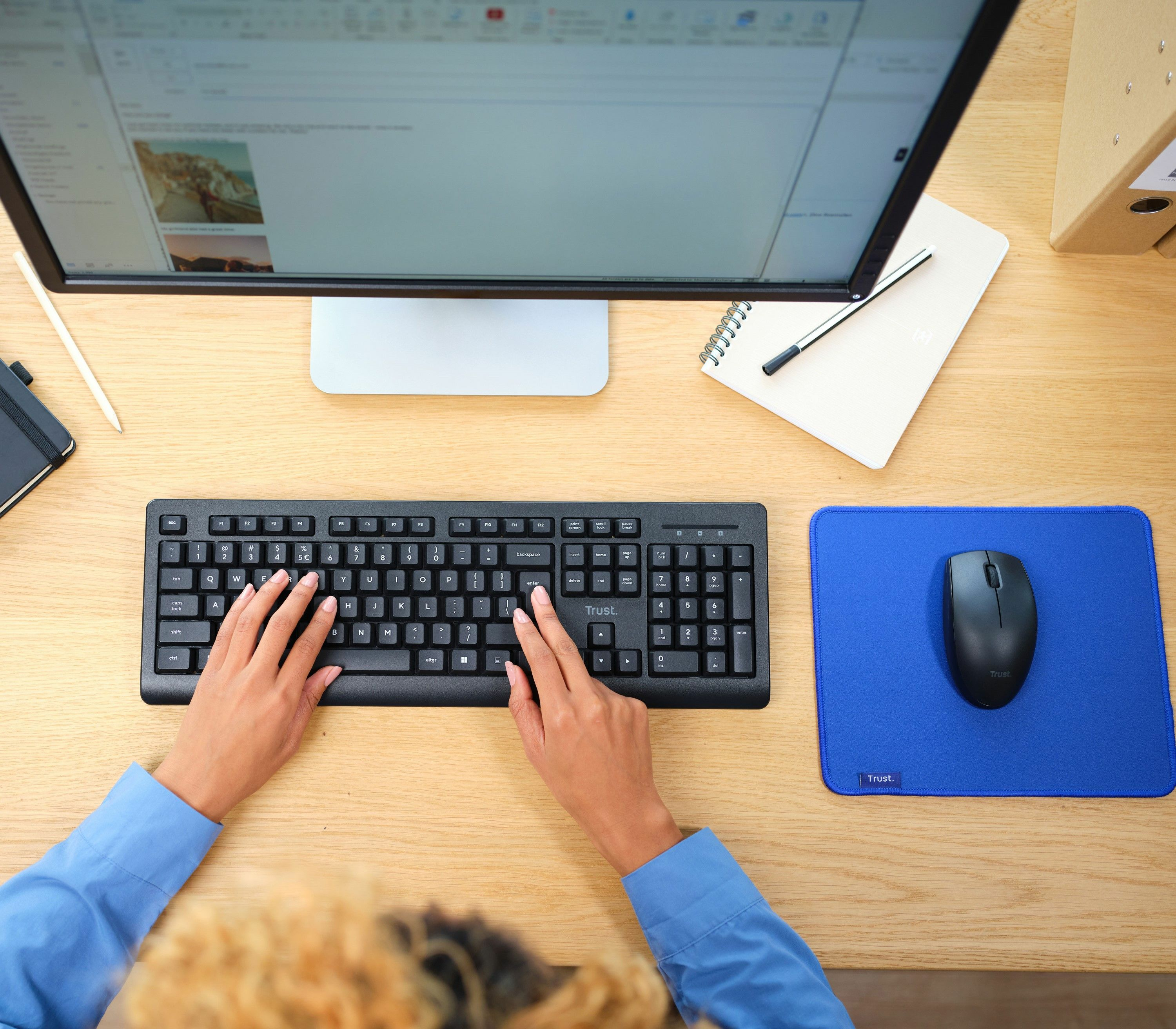 Ręce piszące na czarnej klawiaturze Trust na drewnianym biurku z niebieską podkładką i myszą.