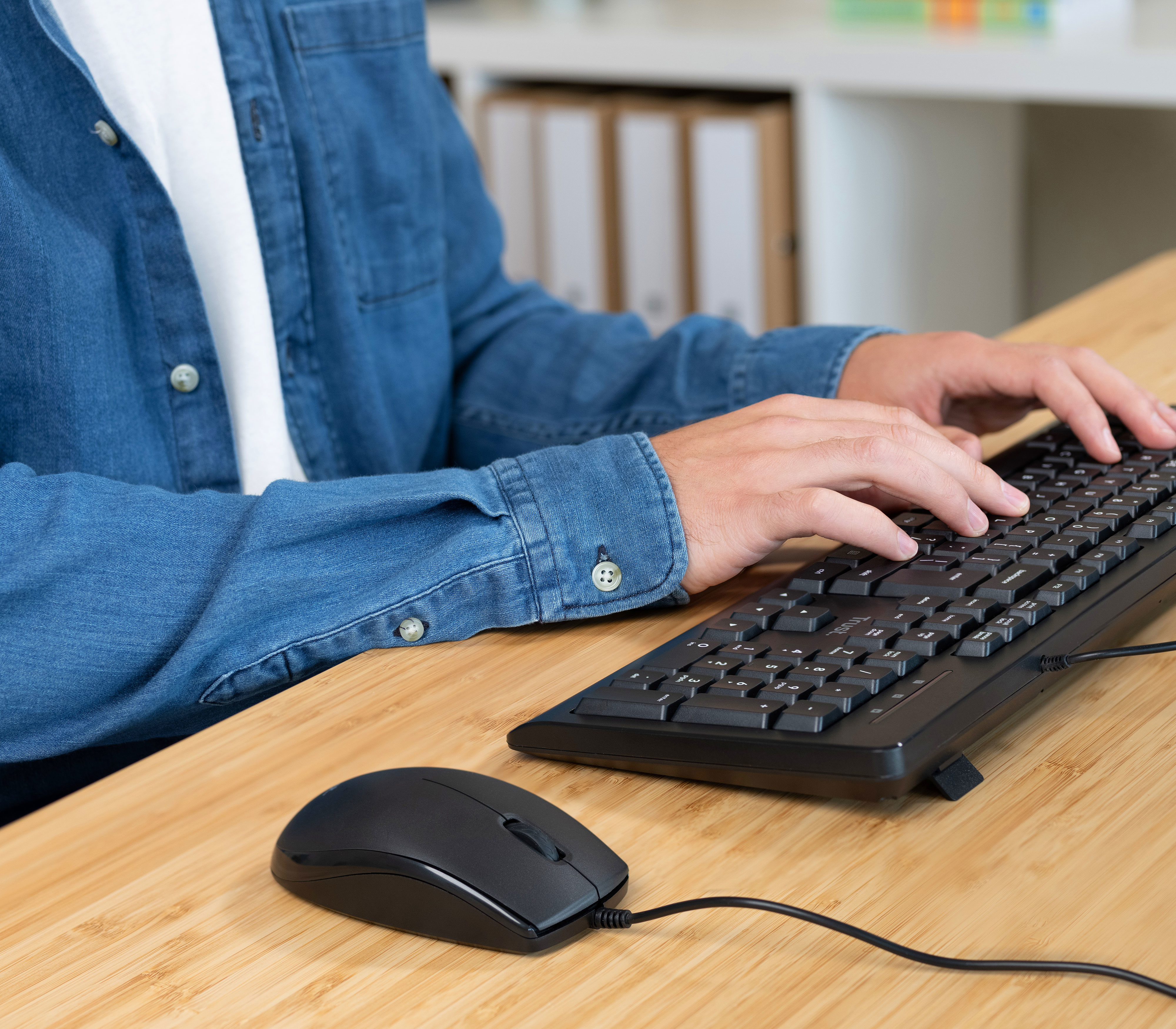 Eine Person benutzt Tastatur und Maus auf einem Holztisch, trägt ein Jeanshemd.
