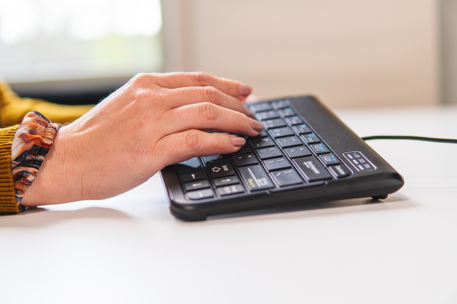 Mano de una persona tecleando en un teclado negro pequeño. El fondo es un escritorio blanco.