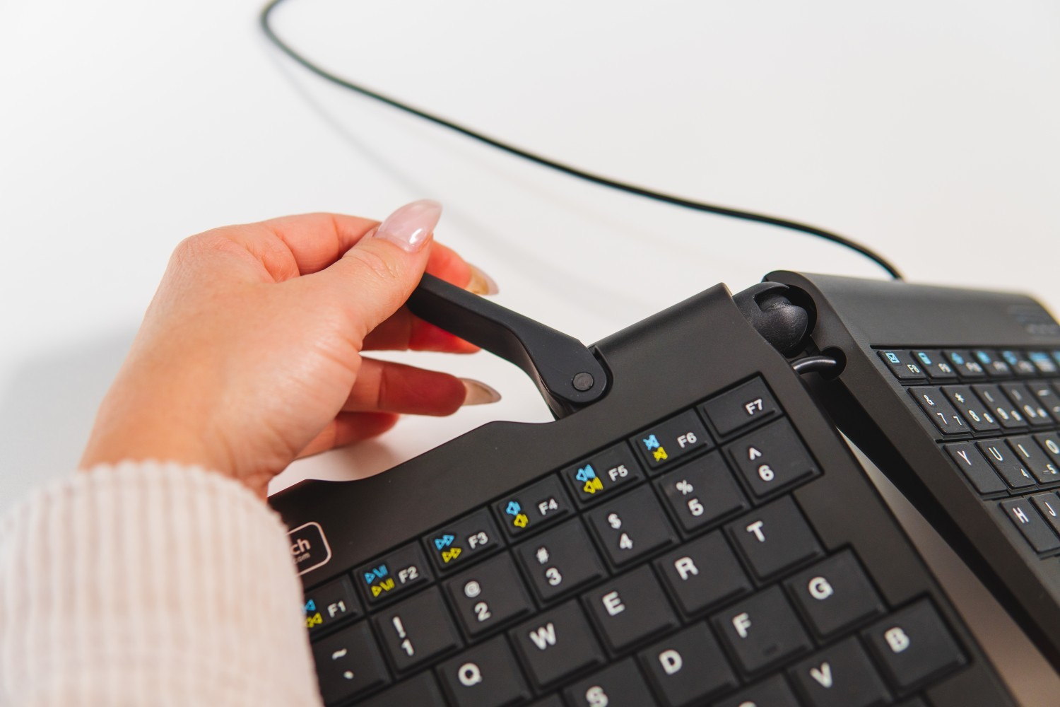 Mano de una persona ajustando un teclado dividido negro sobre una superficie blanca. El teclado está abierto.