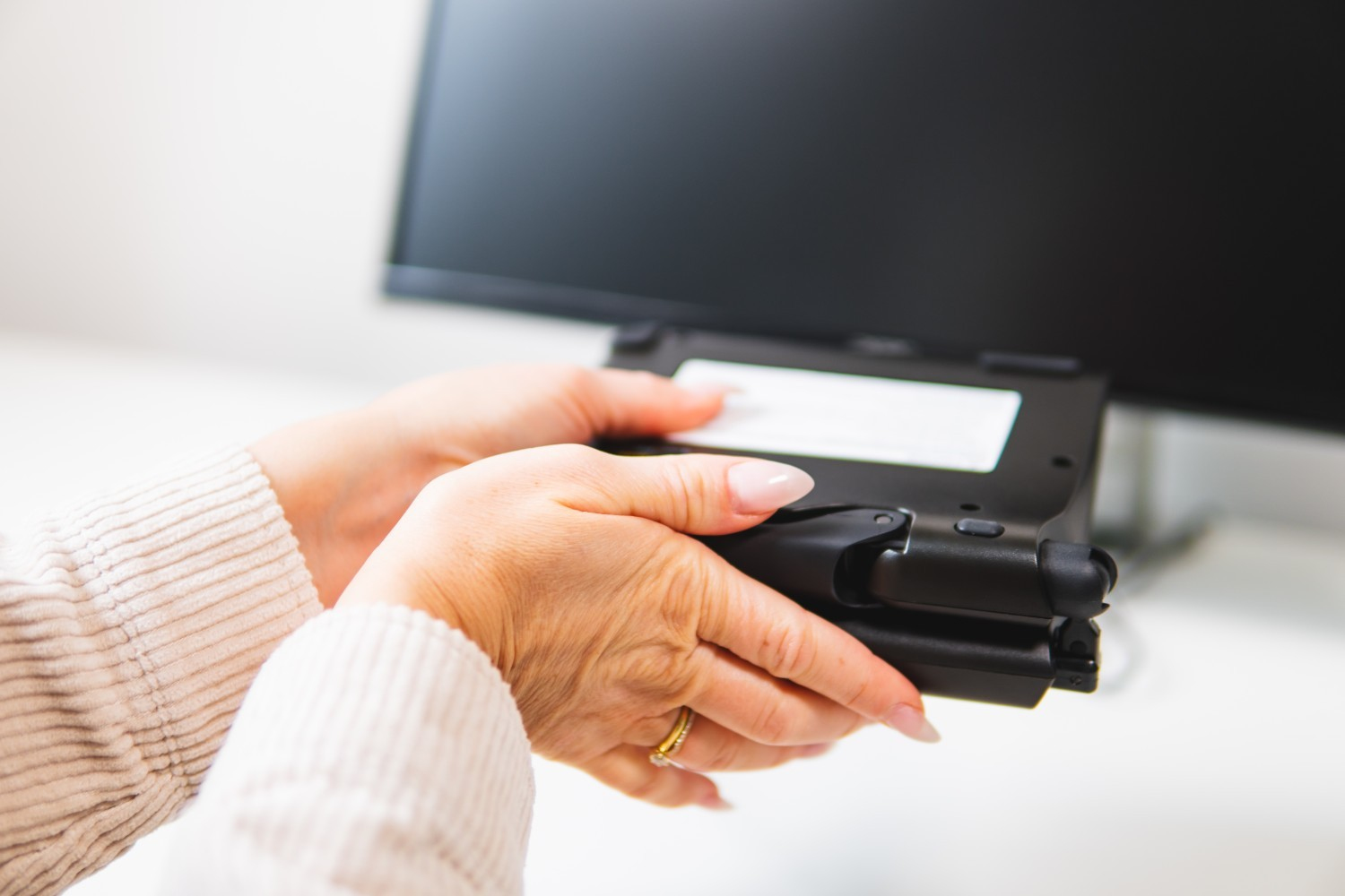 Manos sosteniendo un dispositivo negro frente a un monitor. Escritorio blanco. Enfoque en el dispositivo.
