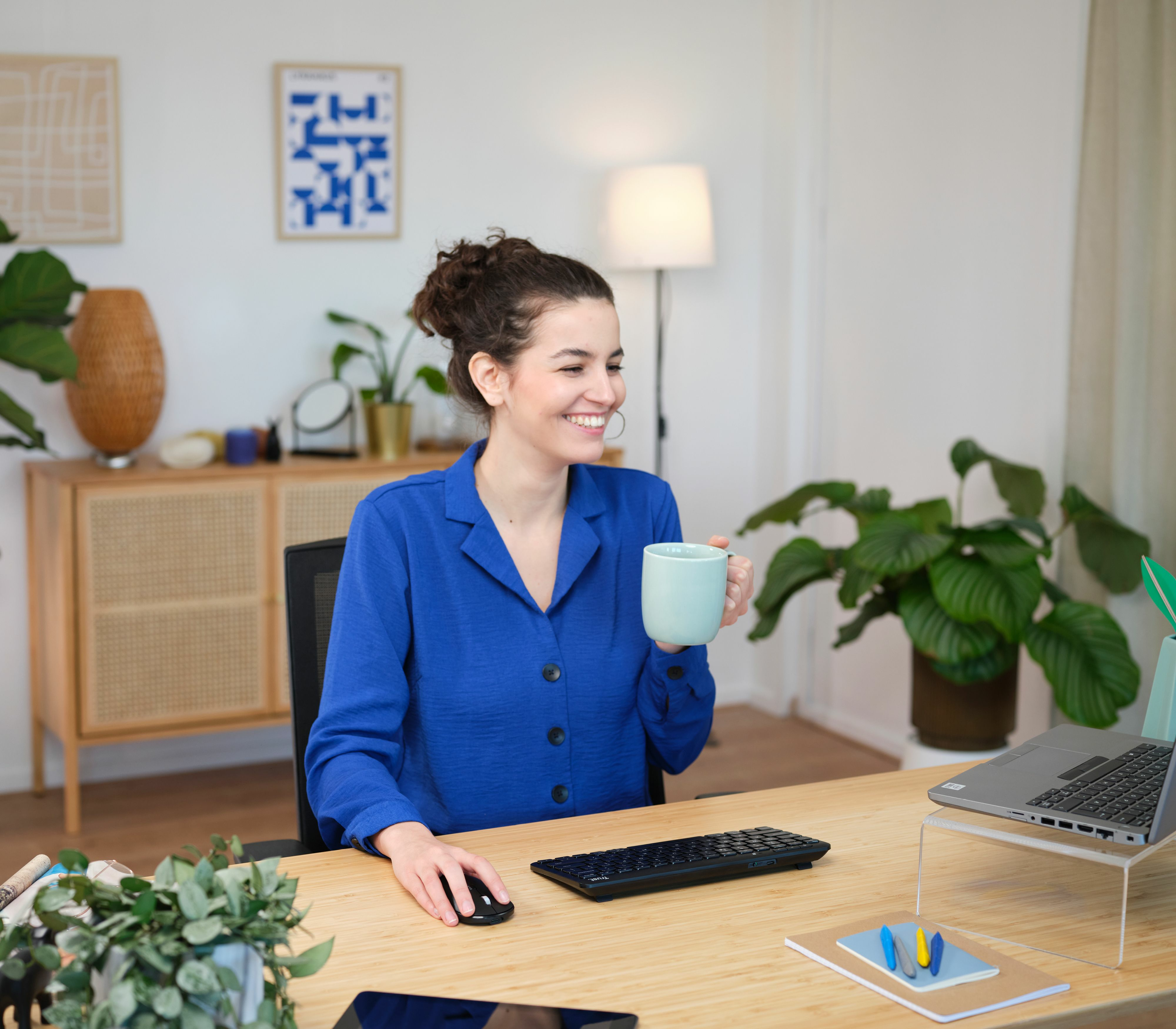Kobieta przy biurku, uśmiechnięta, trzyma kubek, używa klawiatury i myszy.