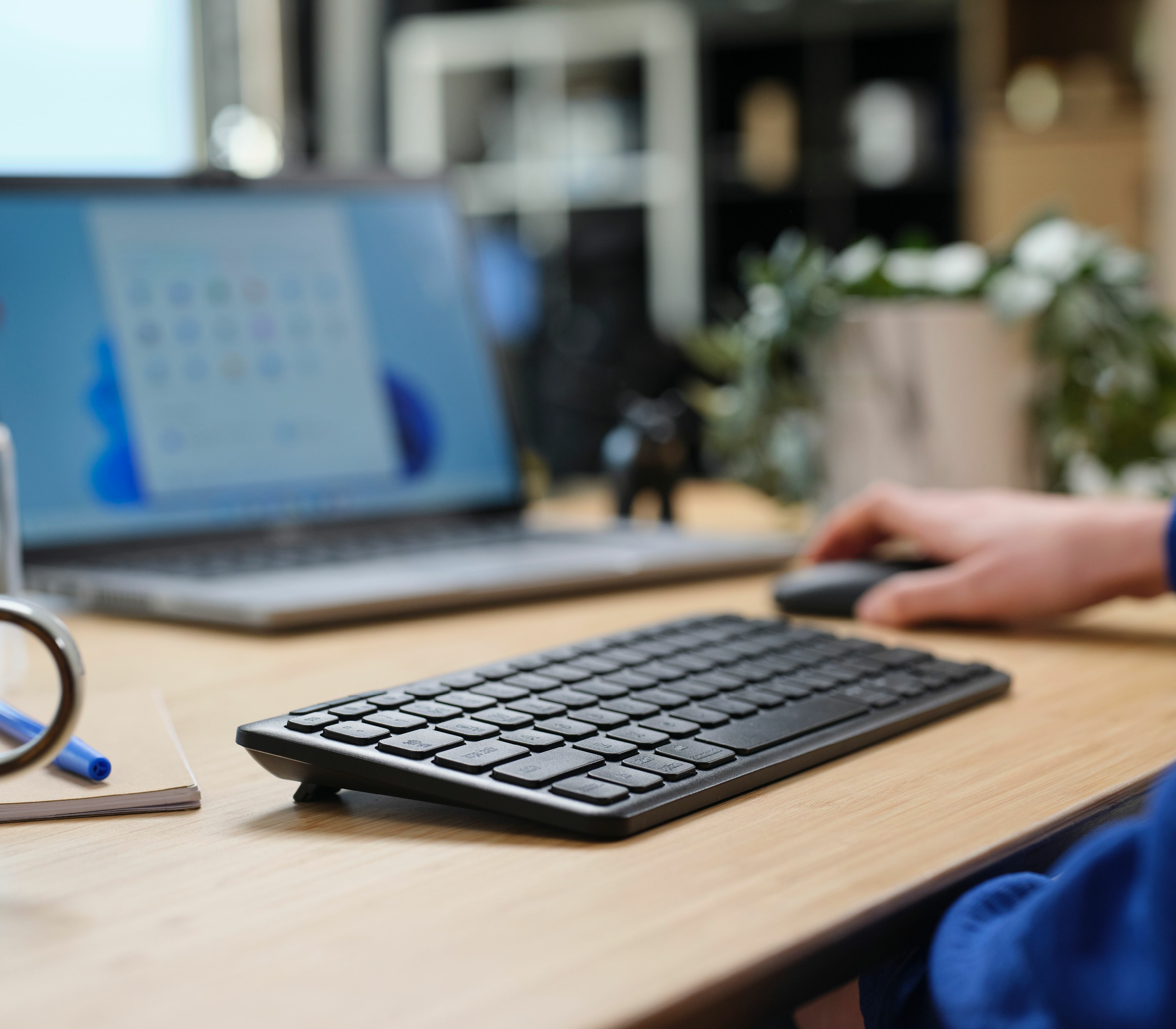 Konfiguracja biurka z laptopem, klawiaturą i myszą. Ręka używa myszy. Biurko jest drewniane.