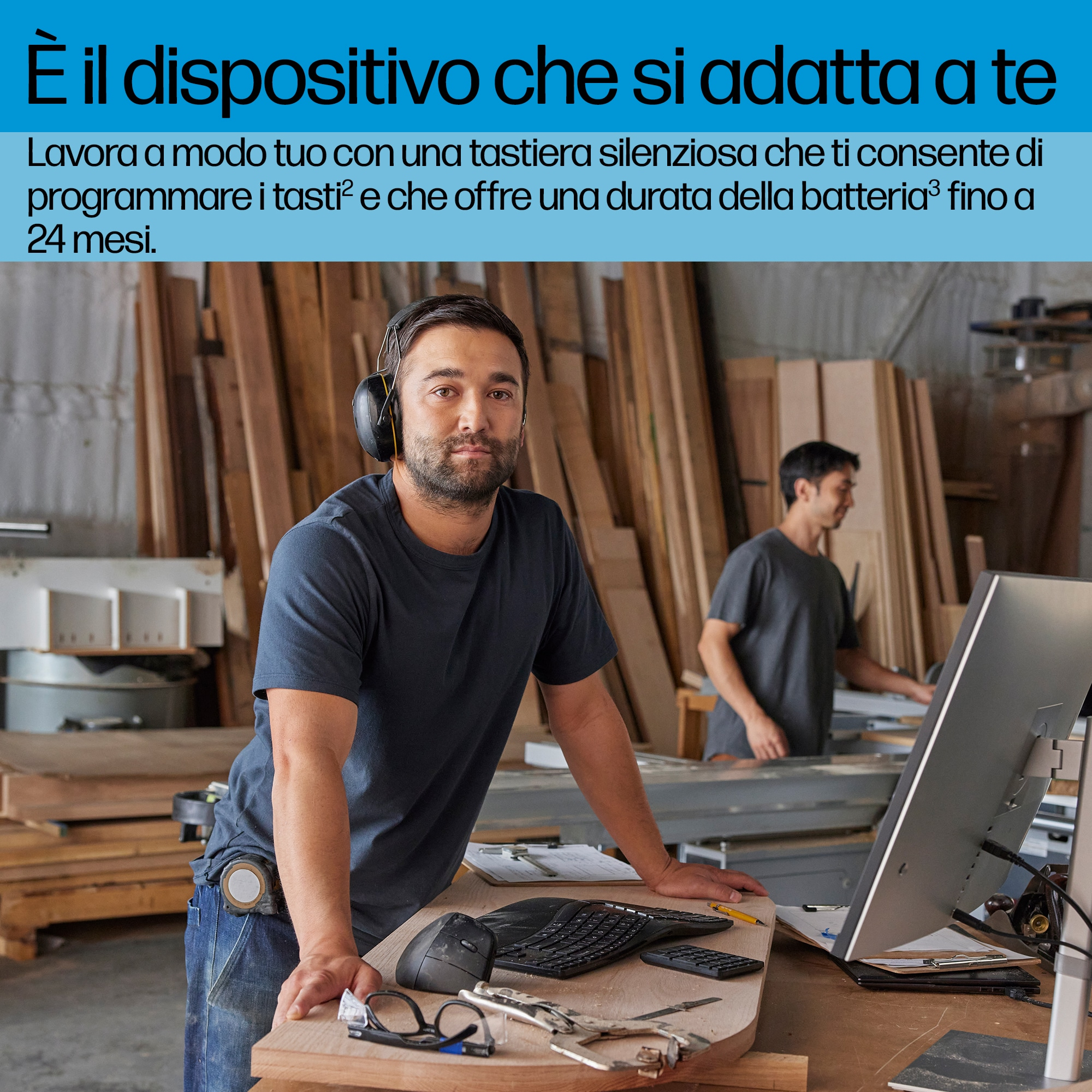 Mężczyzna ze słuchawkami przy biurku z komputerem, klawiaturą i myszą. Drugi mężczyzna w tle.