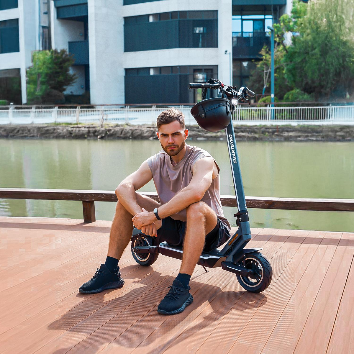 Hombre sentado junto a un scooter eléctrico. Edificio y agua al fondo.