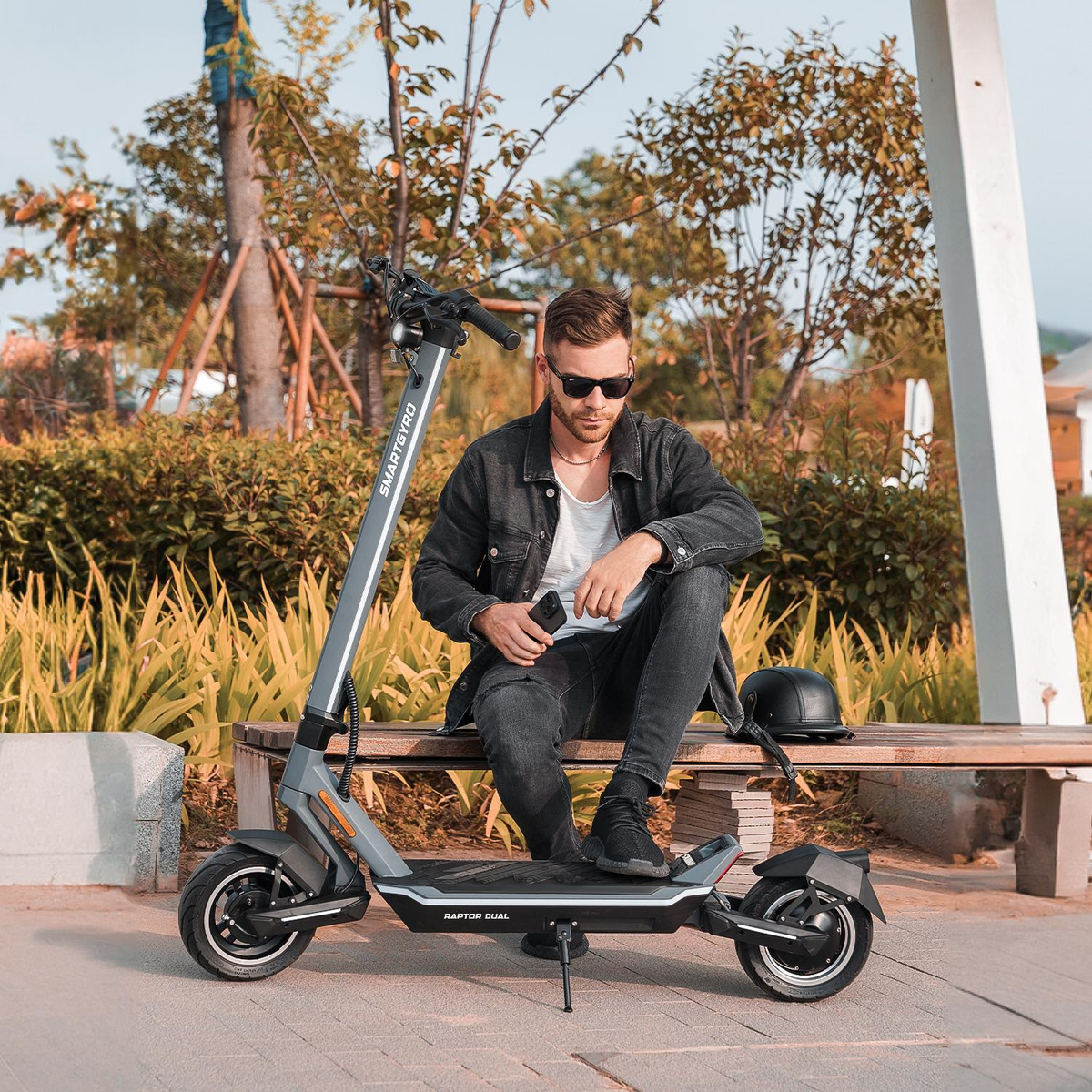 Hombre sentado en un banco con un patinete eléctrico. Lleva gafas de sol y sostiene un teléfono.