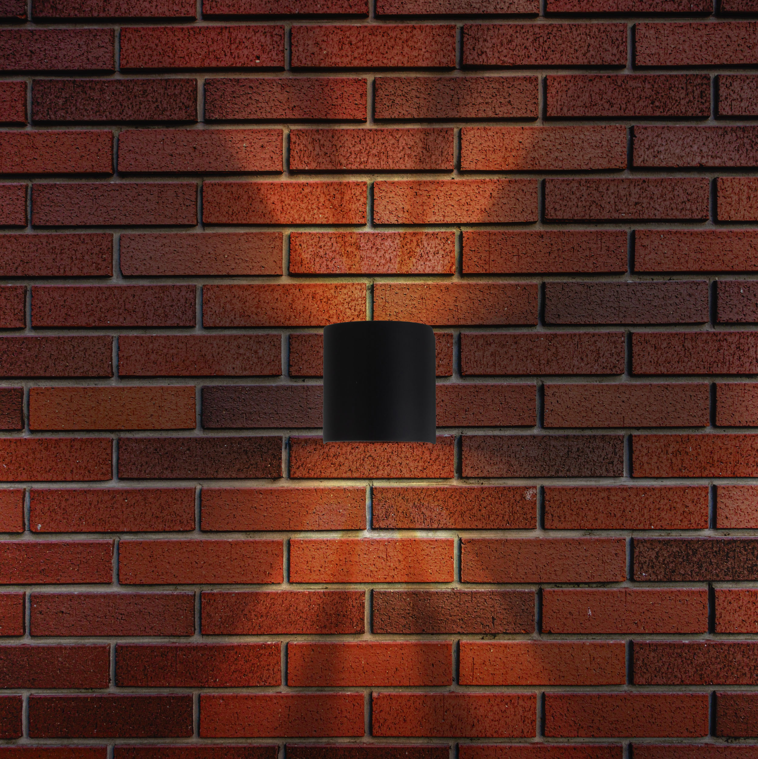Eine schwarze zylindrische Wandleuchte an einer roten Backsteinmauer, von unten beleuchtet.