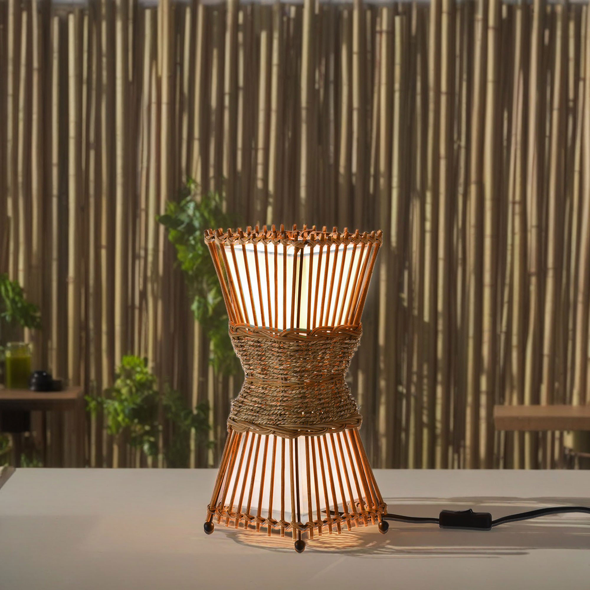 Gewebte braune Lampe mit weißem Innenraum, auf weißer Oberfläche, mit Bambushintergrund und Pflanzen.