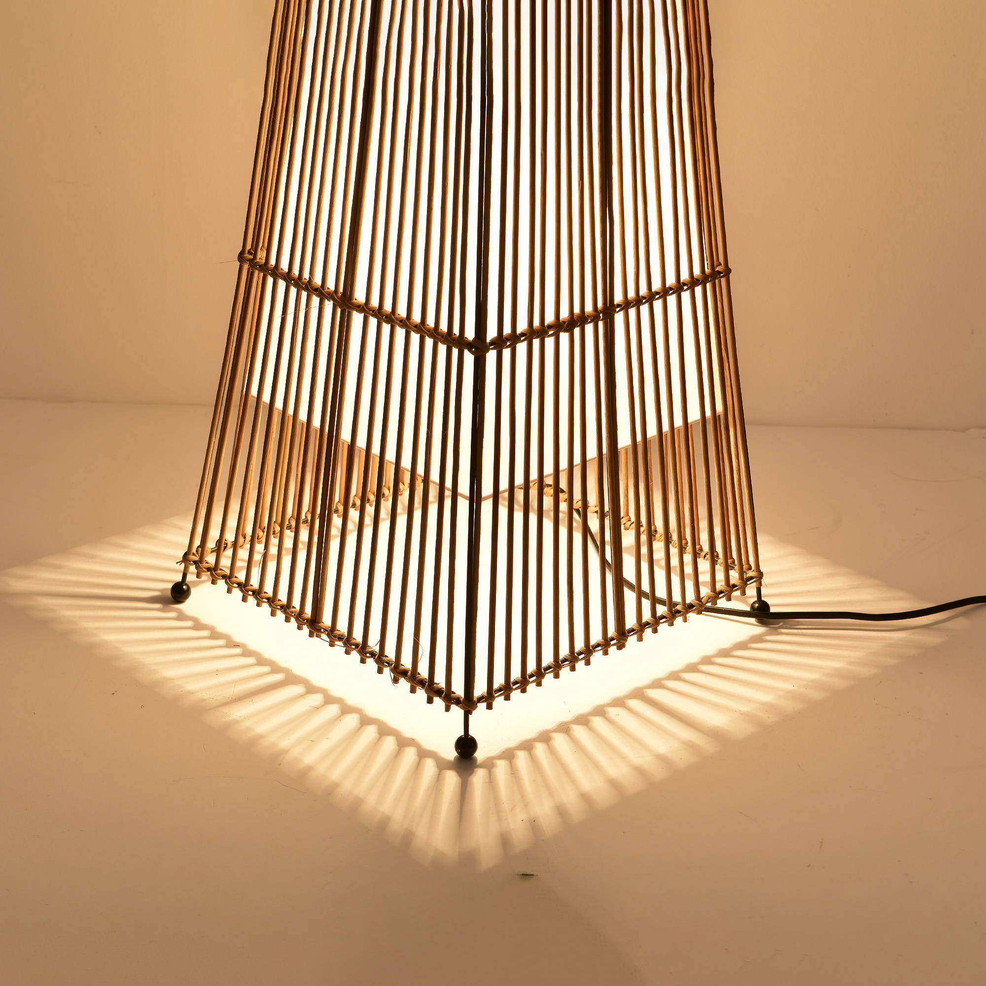 Eine Bambuslampe mit quadratischem Sockel. Die Lampe ist beleuchtet und wirft Schatten auf die weiße Oberfläche.