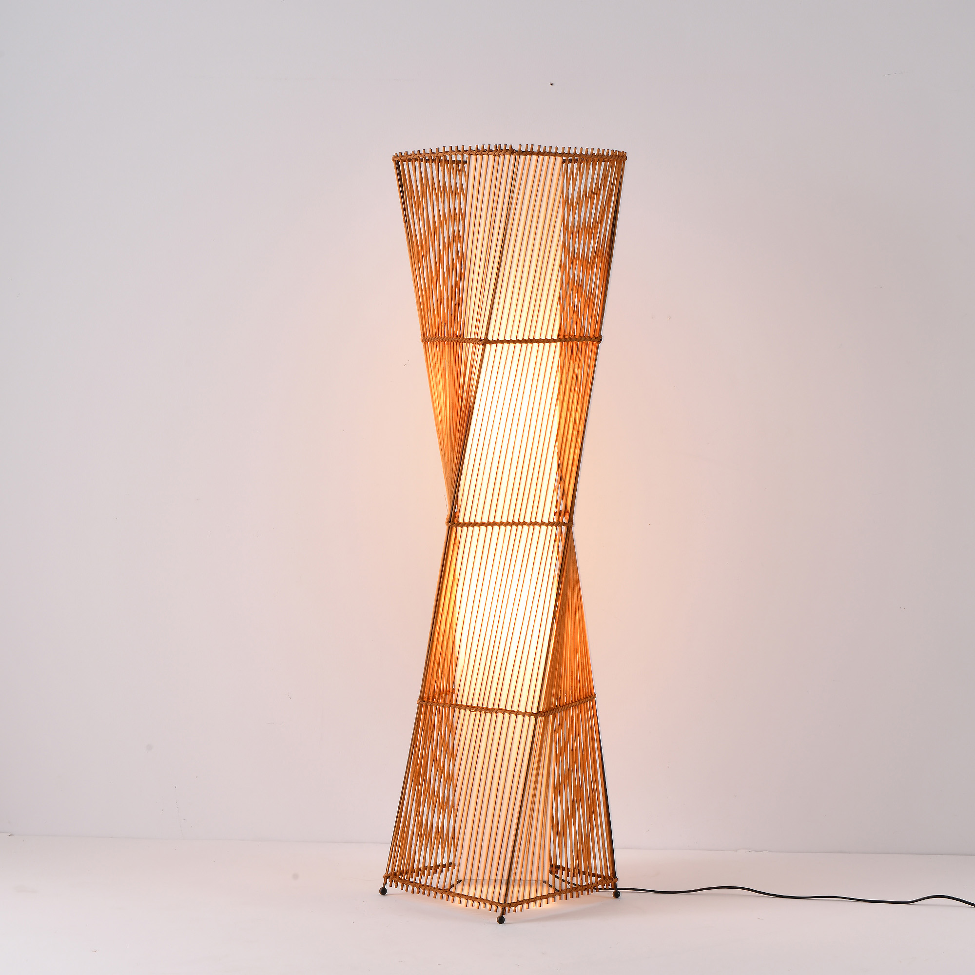 Eine hohe, goldfarbene Stehlampe in einem weißen Raum. Die Lampe besteht aus einem gewebten Material. Das Licht ist an.