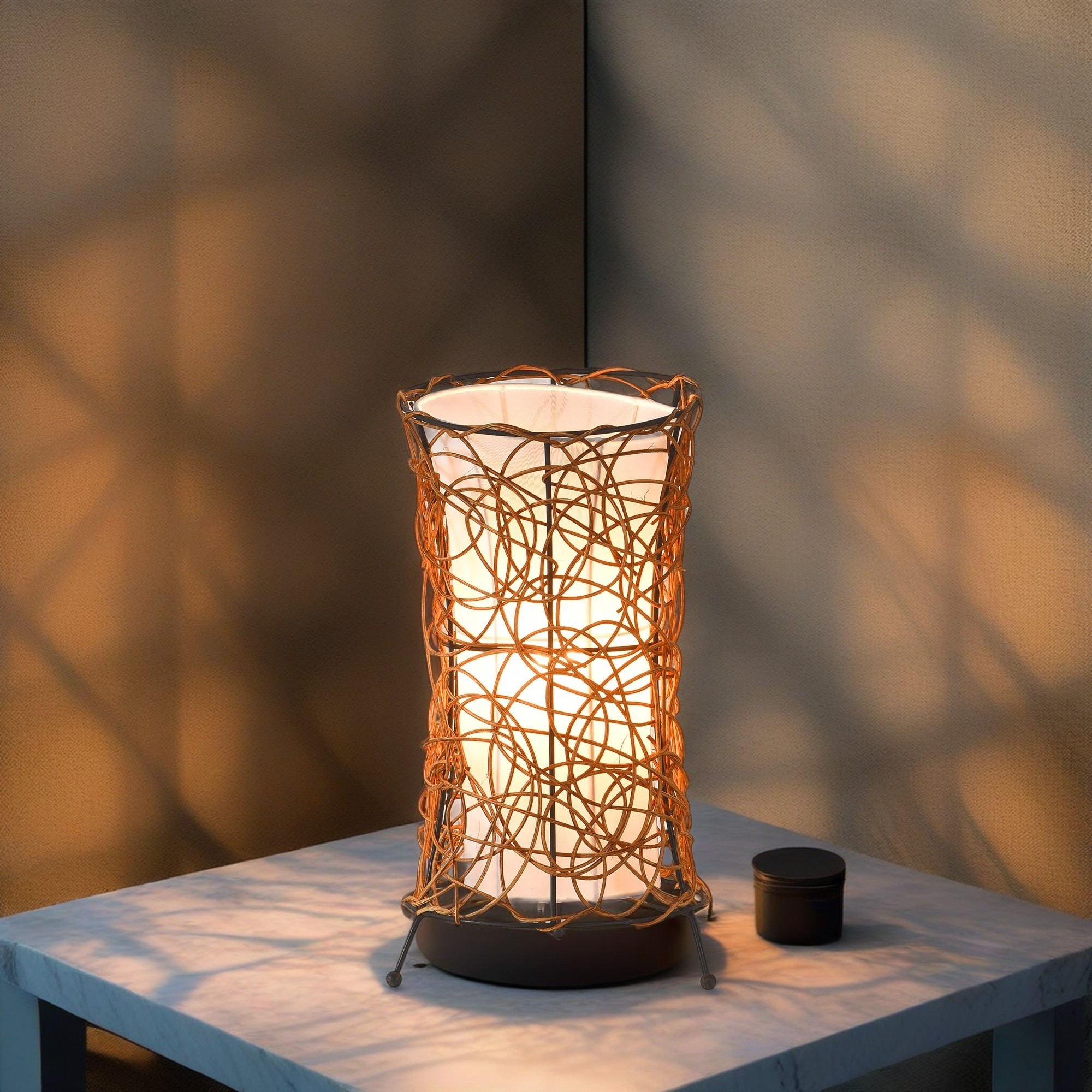 Eine dekorative Lampe mit gewebtem Design. Sie steht auf einem Tisch, daneben ein kleiner schwarzer Behälter.