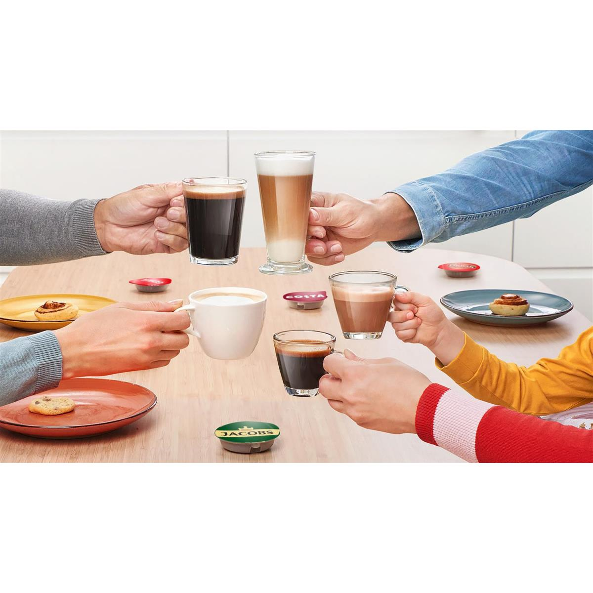 Mani che tengono varie bevande al caffè e capsule di caffè Jacobs, con pasticcini sui piatti.