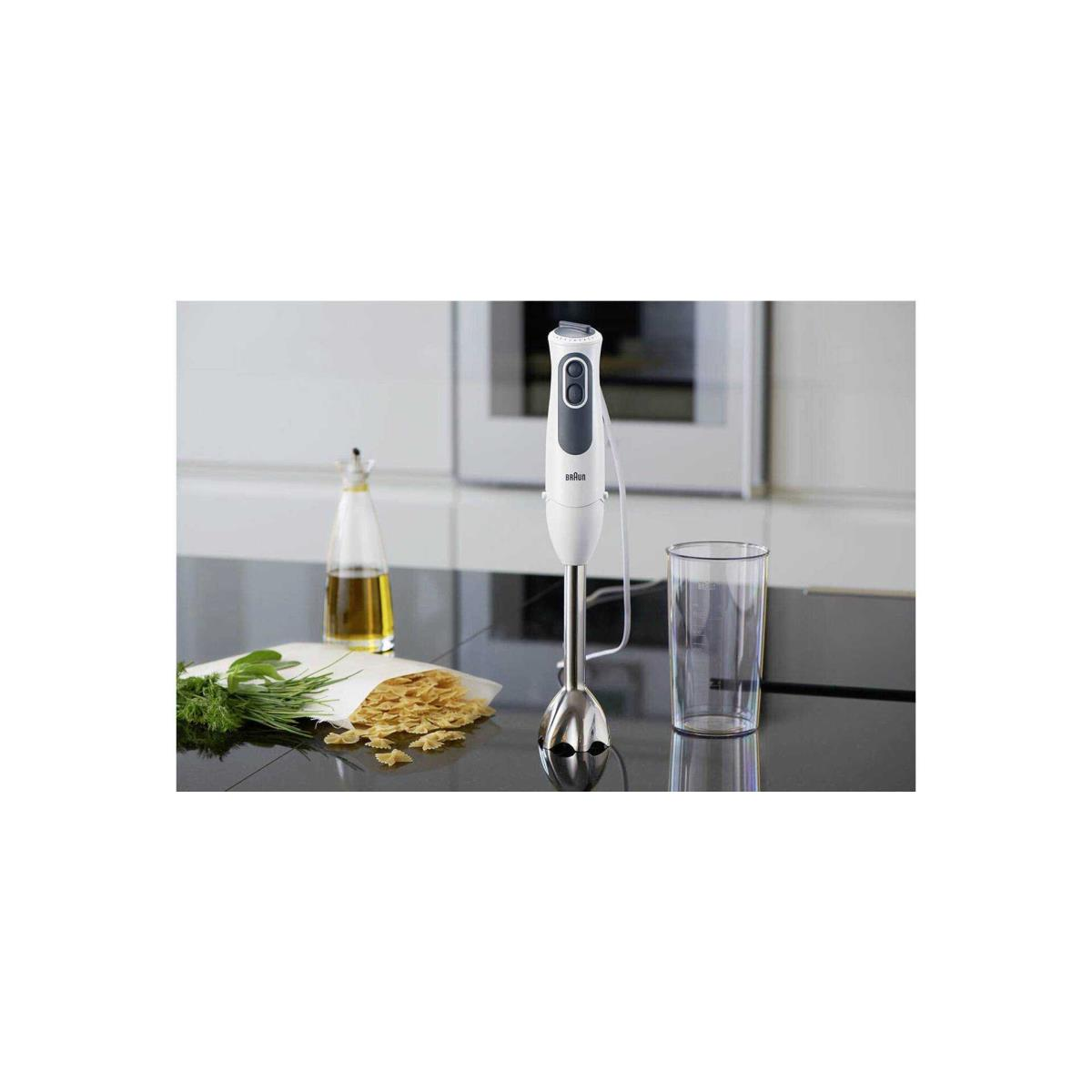 Batidora de mano, blanca con partes plateadas, con alimentos: aceite, pasta y hierbas en superficie negra.