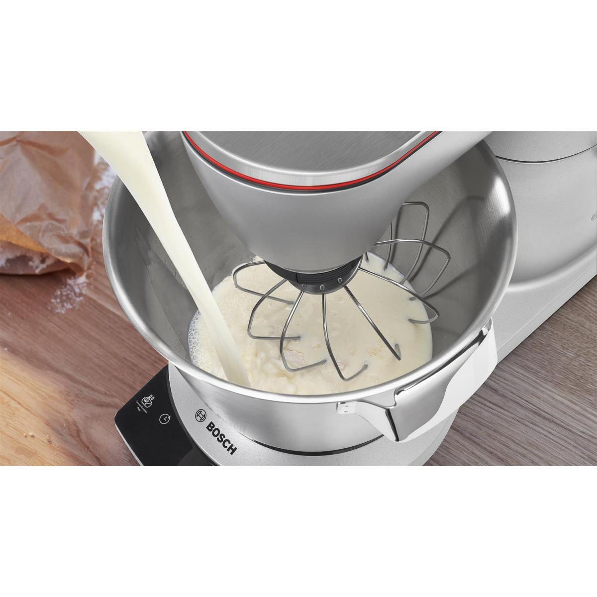 Ein silberner Bosch-Küchenmixer mit Schüssel und Schneebesen, Milch wird in die Schüssel gegossen.