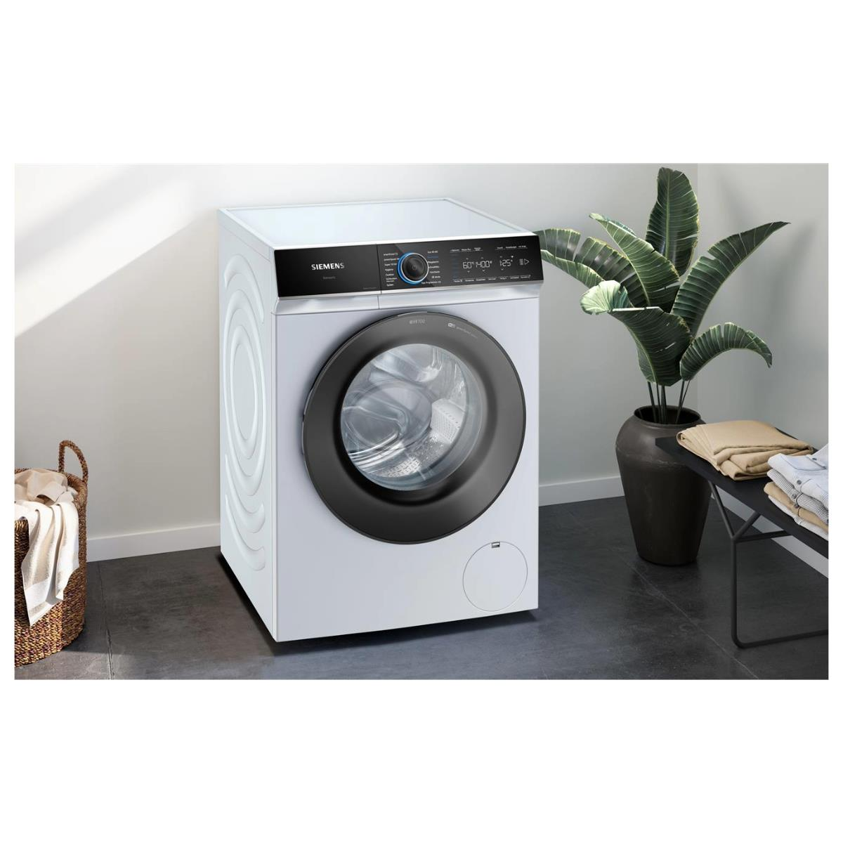 Biała pralka Siemens w dobrze oświetlonym pokoju, z rośliną i koszem w pobliżu.