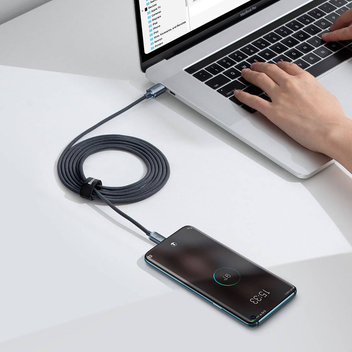 Laptop laadt telefoon op met grijze kabel. Hand op toetsenbord.